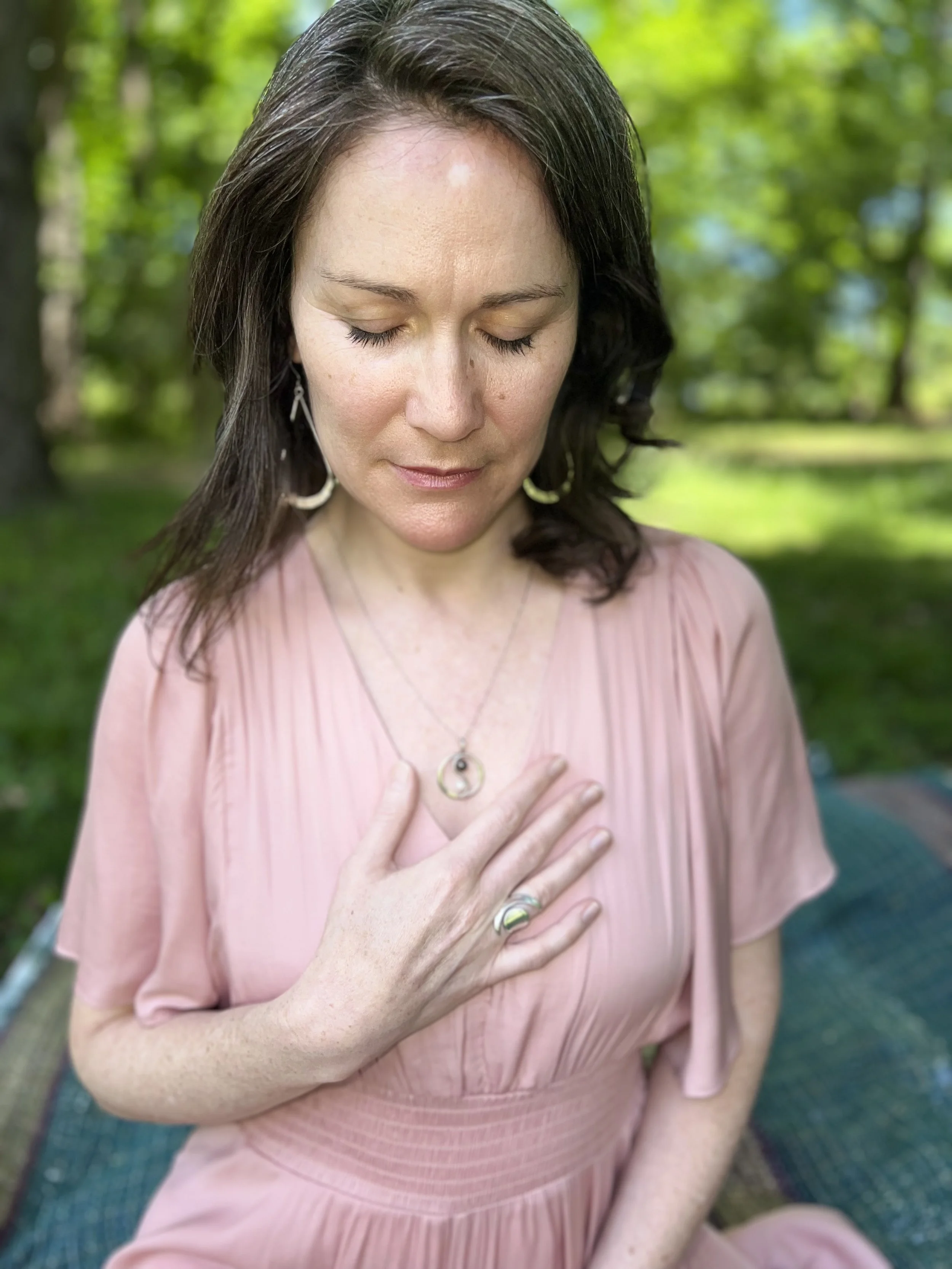 A woman with dark brown hair and light skin wearing a pink dress, with her eyes closed and her left hand resting on her chest, sitting outdoors with green trees and grass in the background.