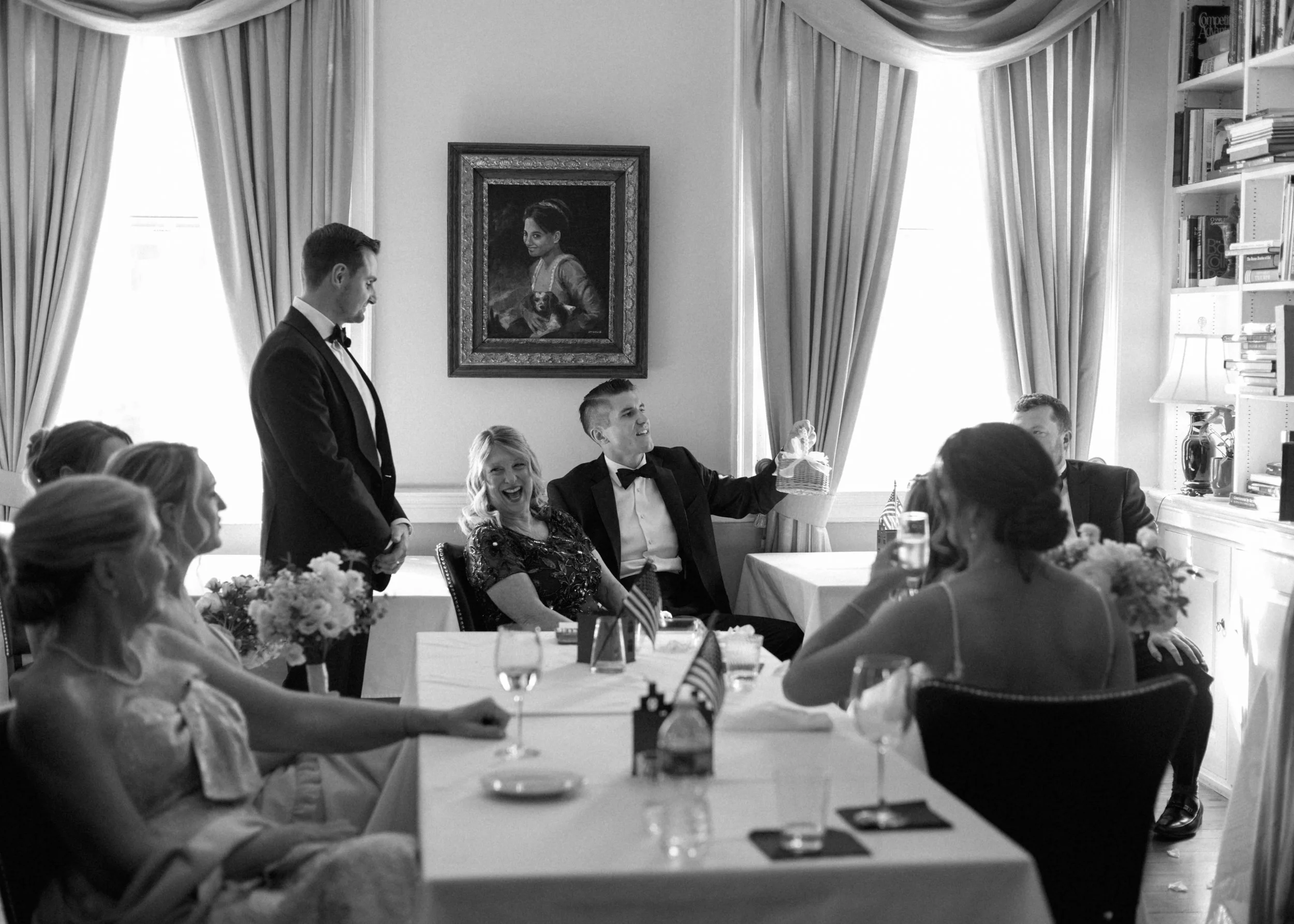 Black and white photo of a formal gathering in a decorated room with large windows and curtains. People in tuxedos and formal dresses sit at tables, some smiling and chatting. A man in a tuxedo stands, while a woman seated at the table is smiling and