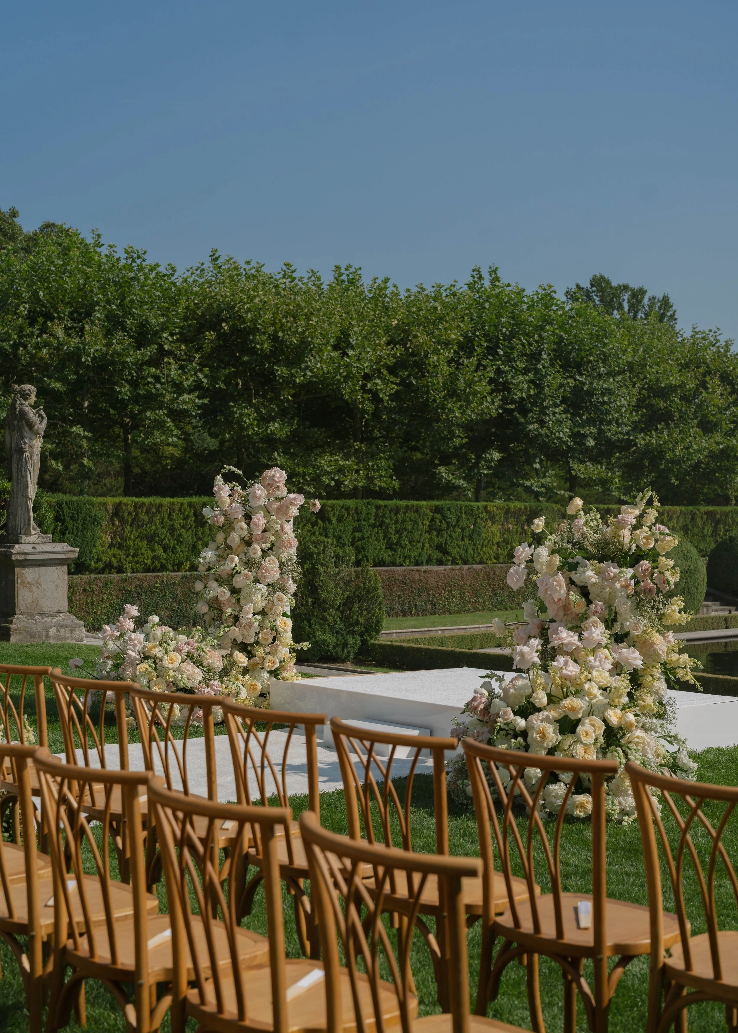 An outdoor wedding setup with wooden chairs arranged in rows facing a decorated altar table with floral arrangements, on a lush green lawn with trees and a blue sky in the background.