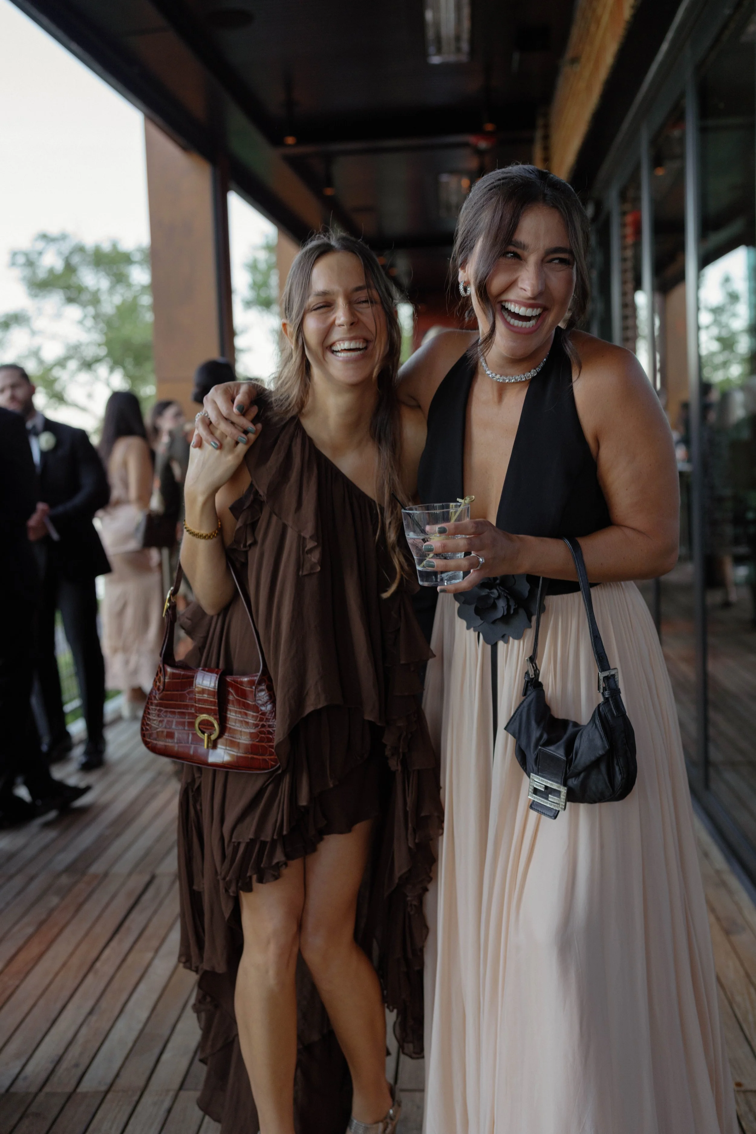 Two women dressed up and laughing at an outdoor event, with one holding a drink in her hand.