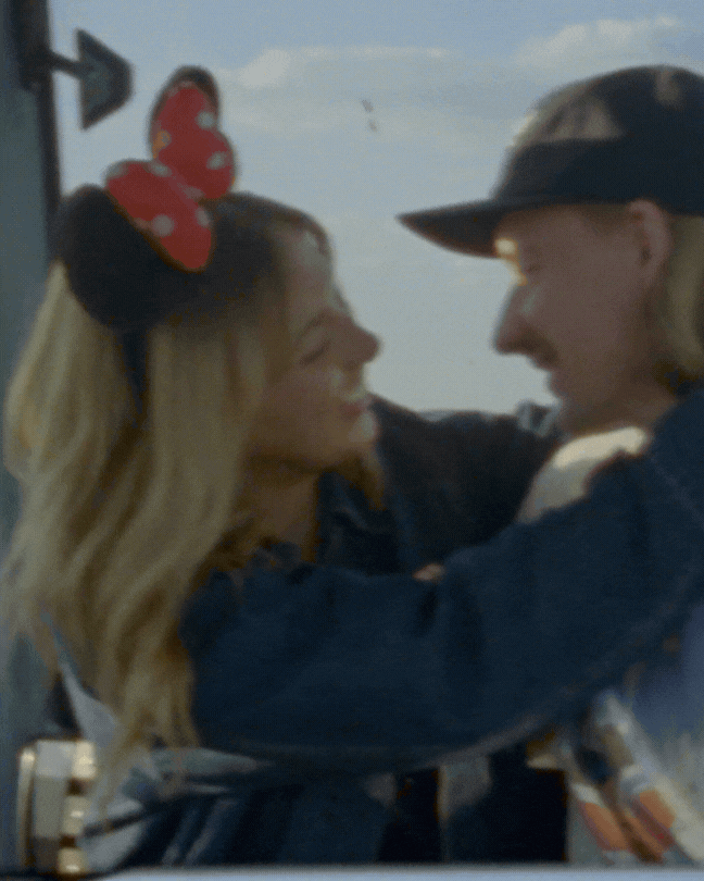 Two people are smiling and looking at each other, embracing with their arms around each other's shoulders. The woman has blonde hair with a red bow headband, the man is wearing a large black hat. The background shows a cloudy sky.