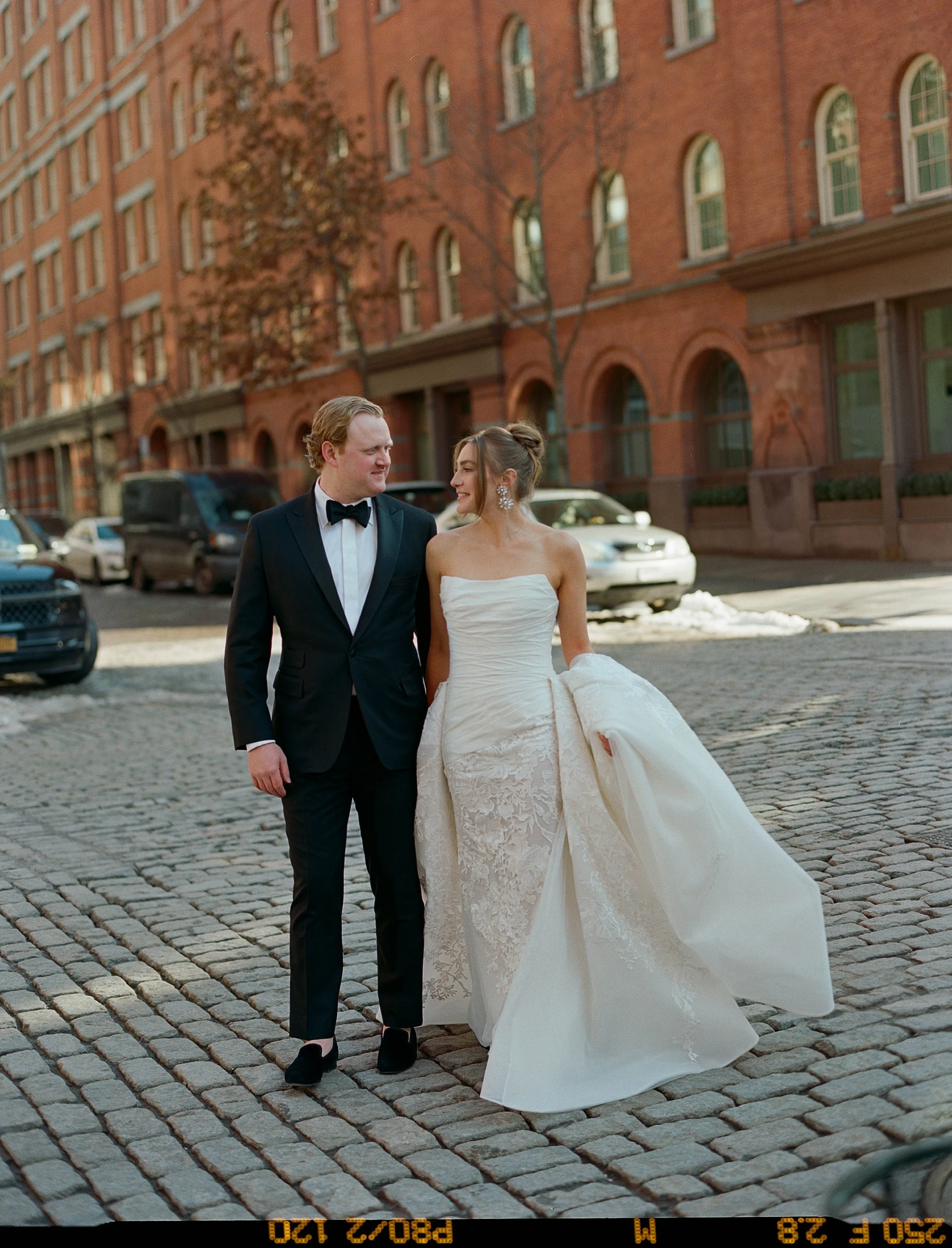 A couple dressed in wedding attire, walking happily on a cobblestone street in front of red brick buildings, with some cars parked along the street and leafless trees in the background.