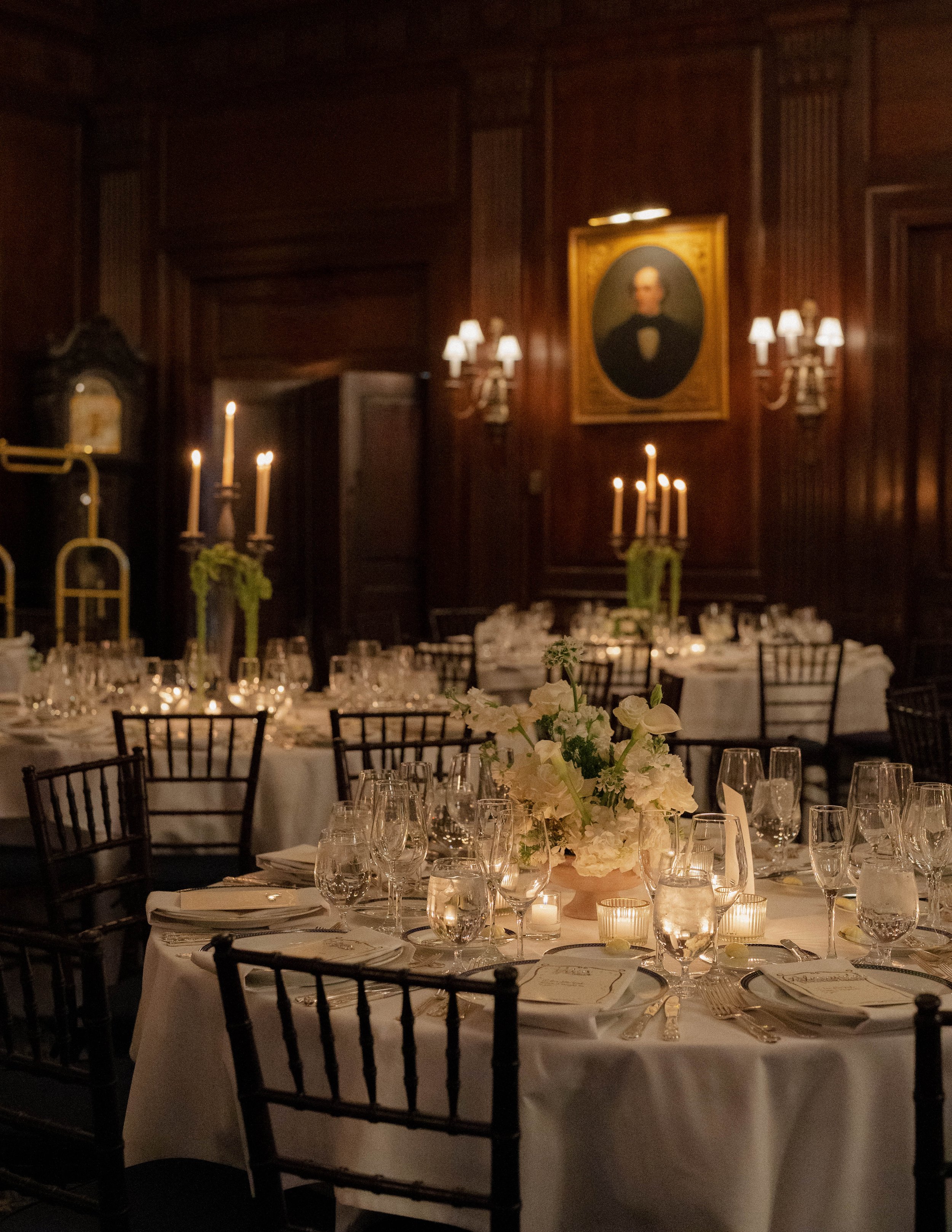 Elegant banquet hall with tables set for a formal event, featuring white tablecloths, floral centerpieces, multiple glasses, and lit candles, with wooden paneling sides, portraits, and wall sconces with lamps.
