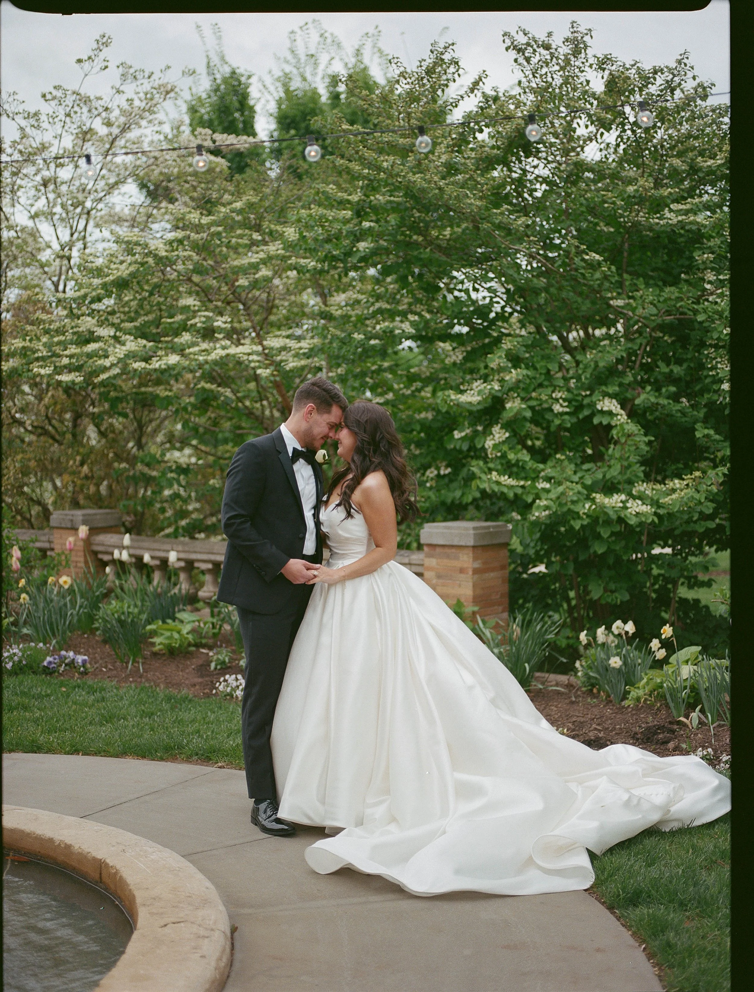 A bride and groom standing closely in a garden, touching foreheads and smiling, with trees and blooming flowers in the background, under string lights hanging overhead.