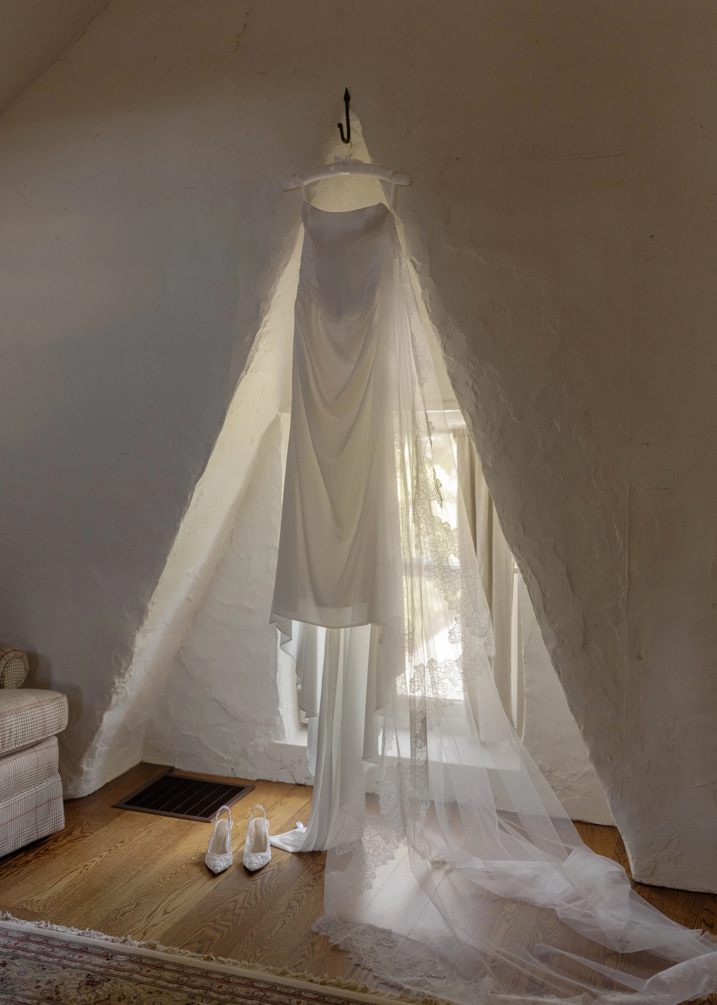 A white wedding dress hangs from a hook on a sloped wall in a cozy room with a window and sheer lace curtains. White wedding shoes are placed on the wooden floor underneath the dress.