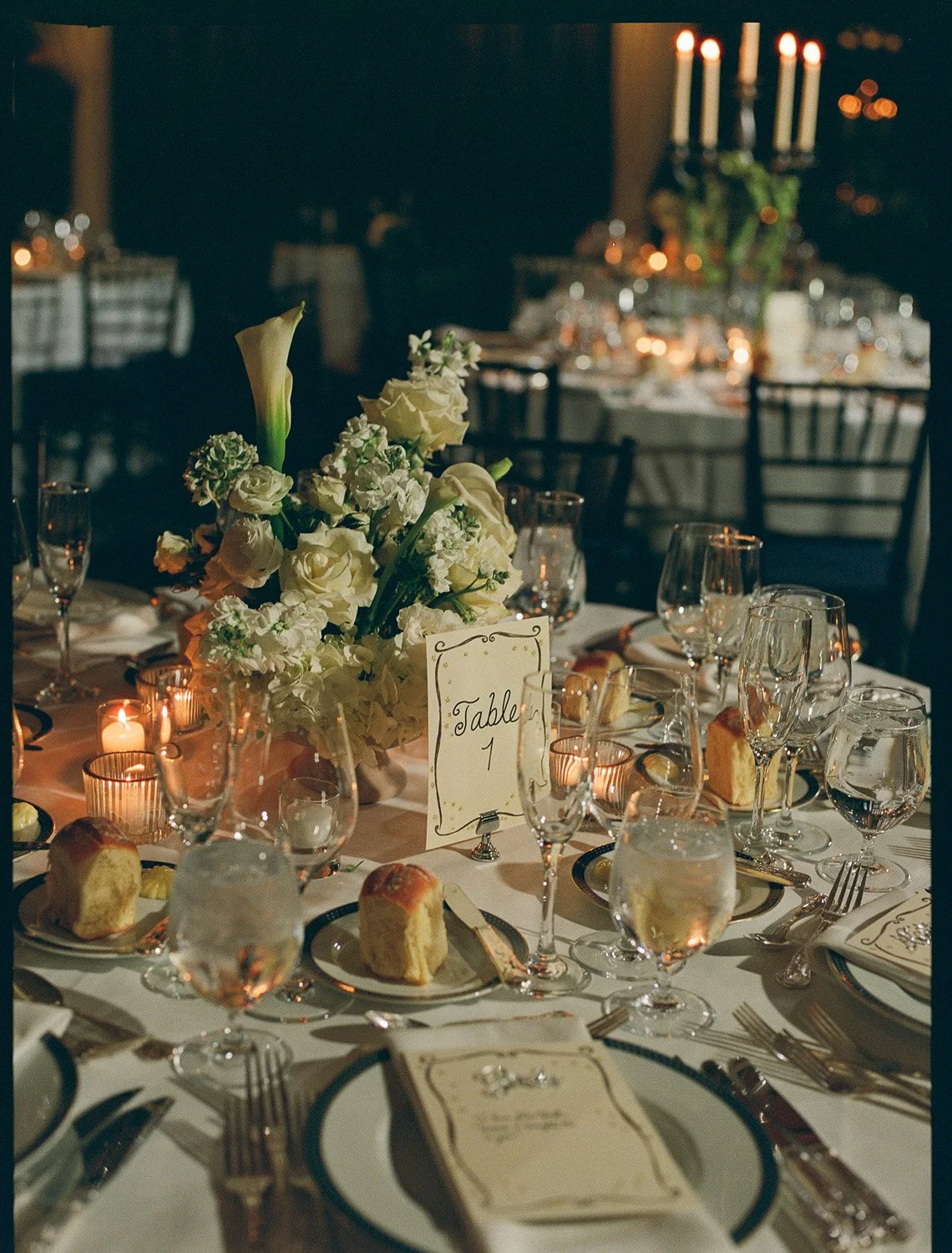 Elegant banquet table set for a formal event with floral centerpiece, candles, glassware, plates, bread rolls, and a table number sign.
