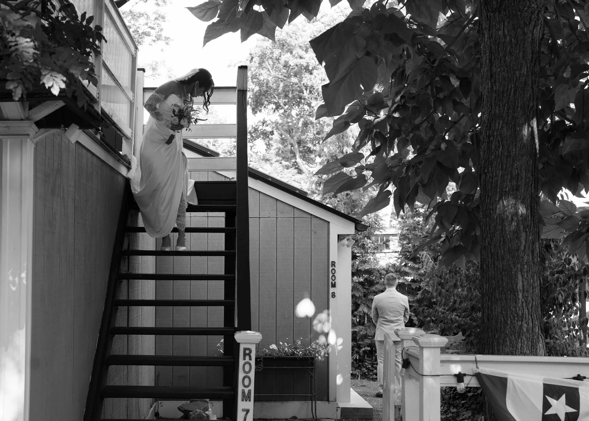 A bride in a wedding dress holding a bouquet stands on the stairway of a small building outdoors, with two men in suits standing nearby. Large trees and leaves are visible in the background.