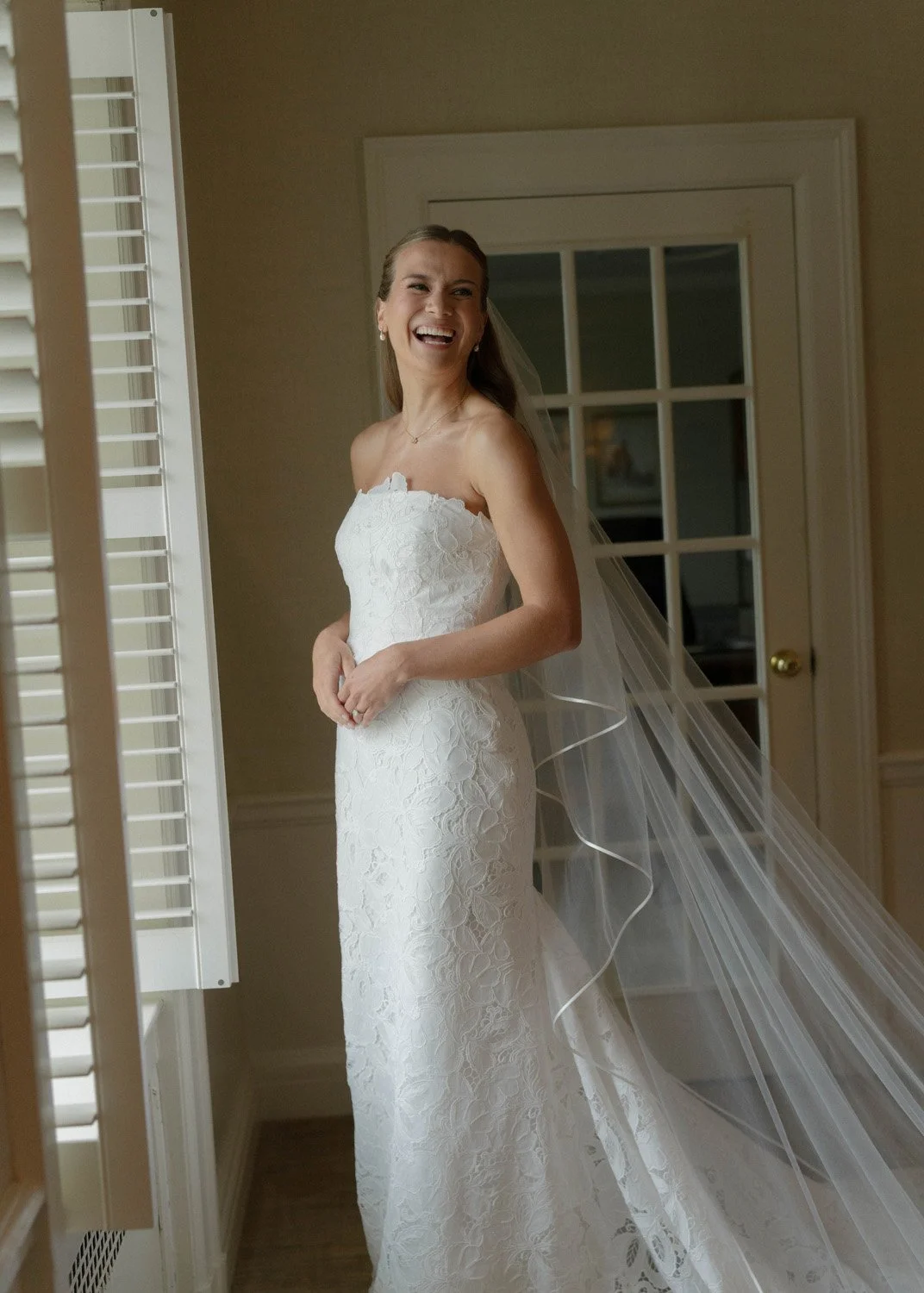 A bride in a strapless white lace wedding dress and veil smiling indoors near a window with white shutters.