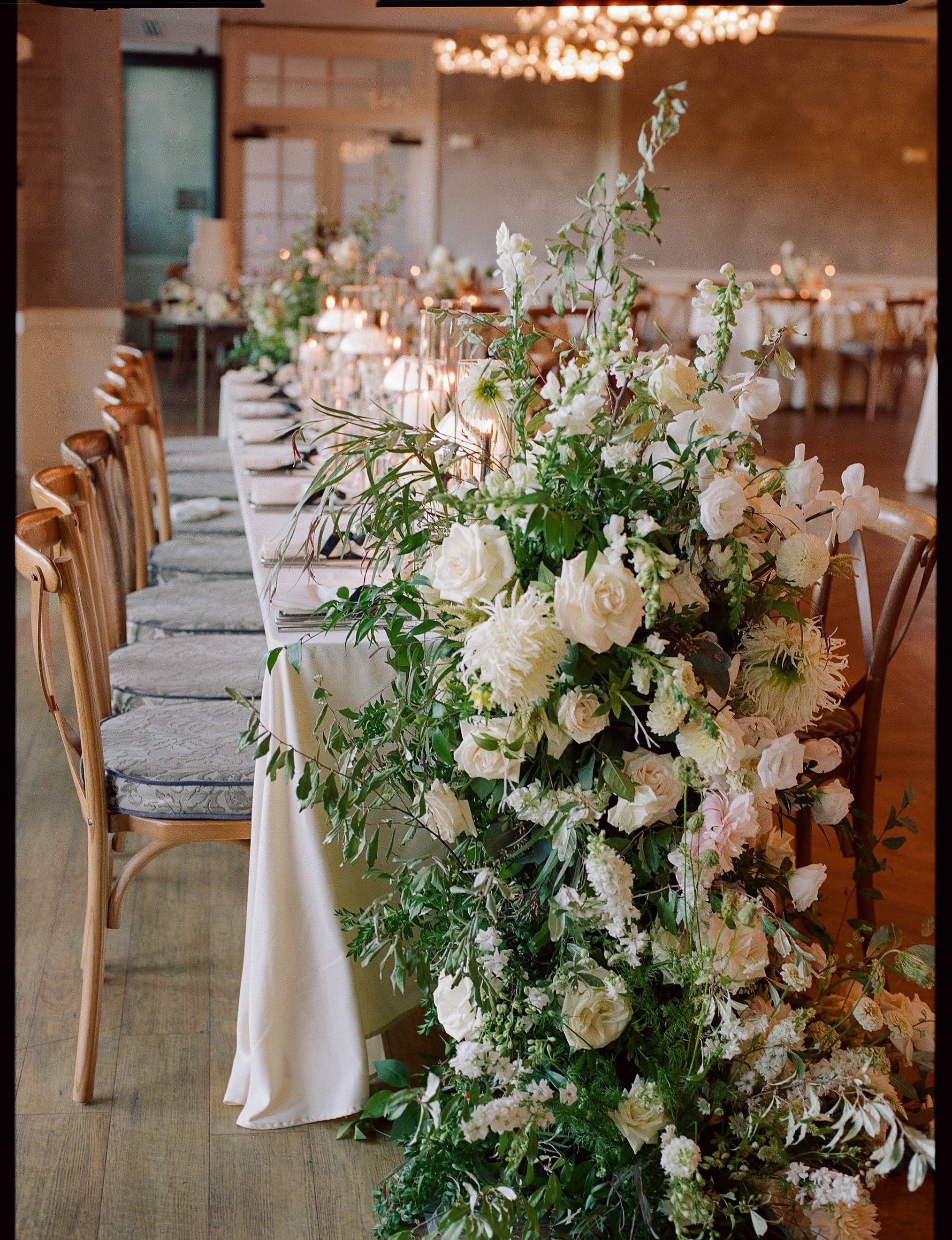 A long dining table decorated with a lush arrangement of white flowers and greenery, set in a well-lit, elegant room.