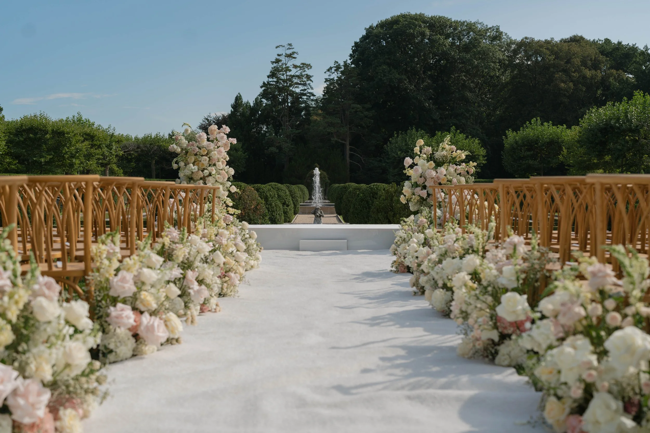 Wedding aisle decorated with white and blush pink flowers on both sides, with wooden chairs and a fountain in the background, under a clear blue sky.