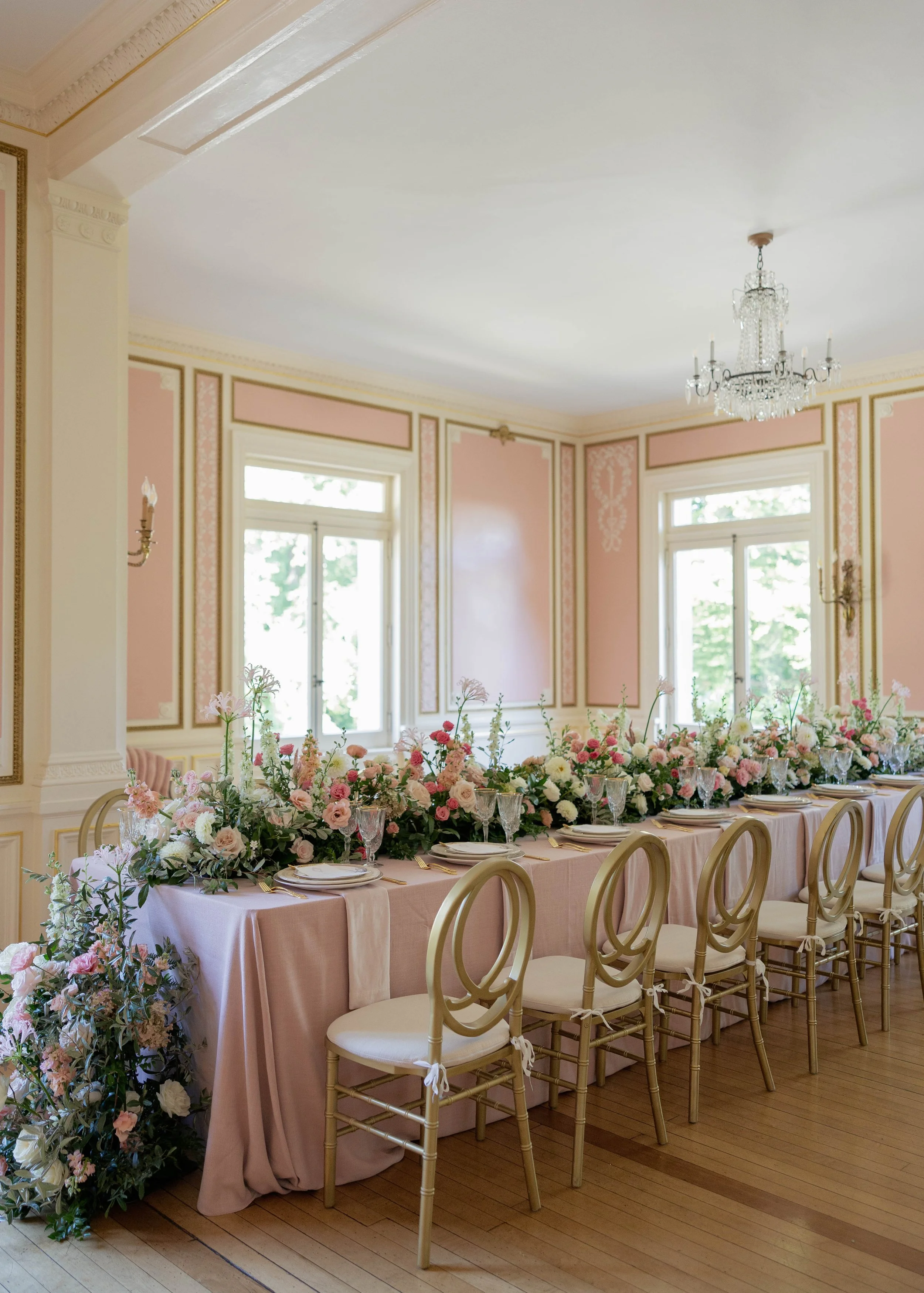 Elegant dining room with pink and gold wall decor, large windows, a crystal chandelier, and a long table decorated with a pink tablecloth and a lush floral centerpiece, surrounded by gold chairs with white cushions.