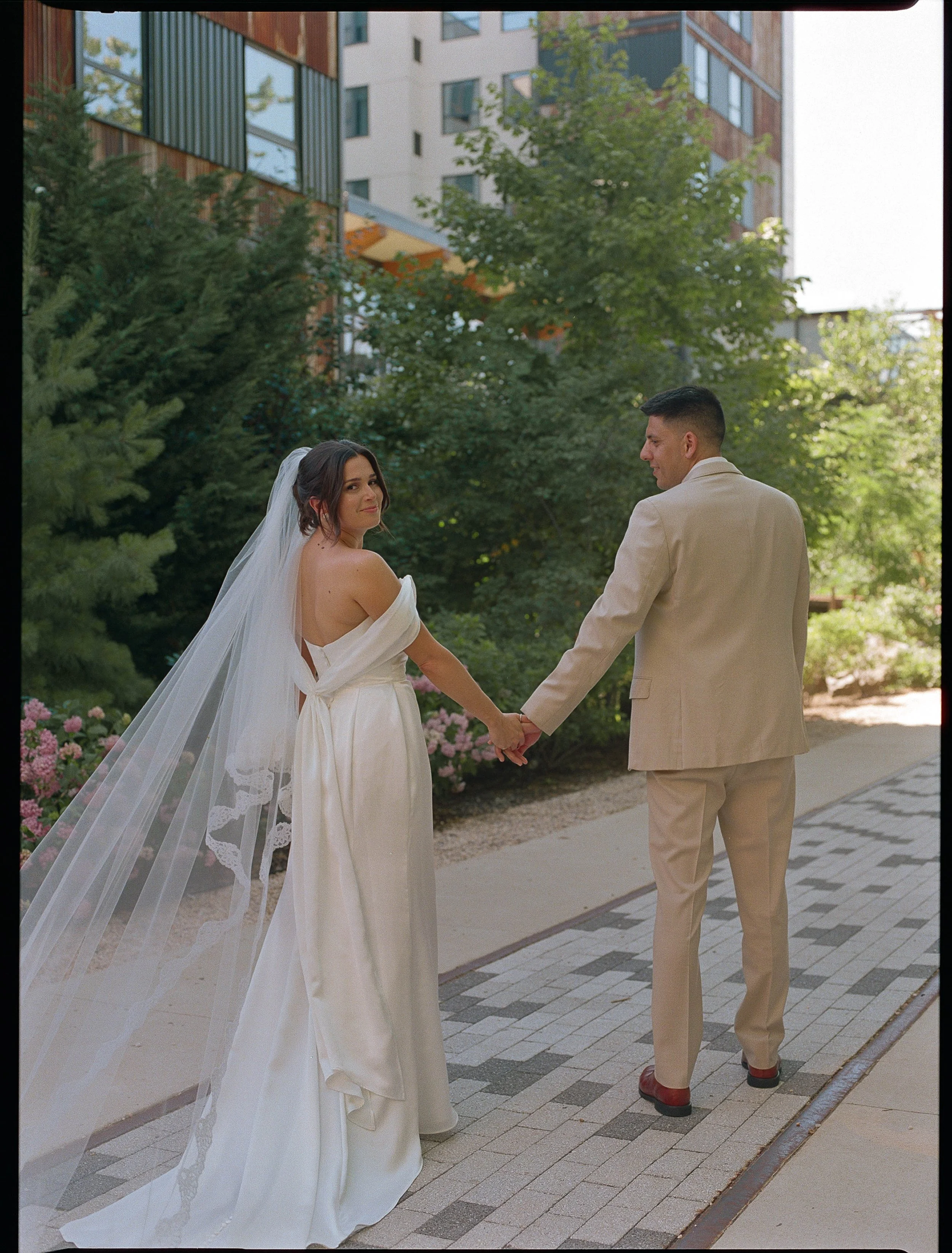 A bride and groom holding hands outdoors on a sunny day. The bride is wearing a white off-shoulder wedding dress with a veil, and the groom is in a beige suit. They are standing on a paved pathway with trees and bushes in the background.