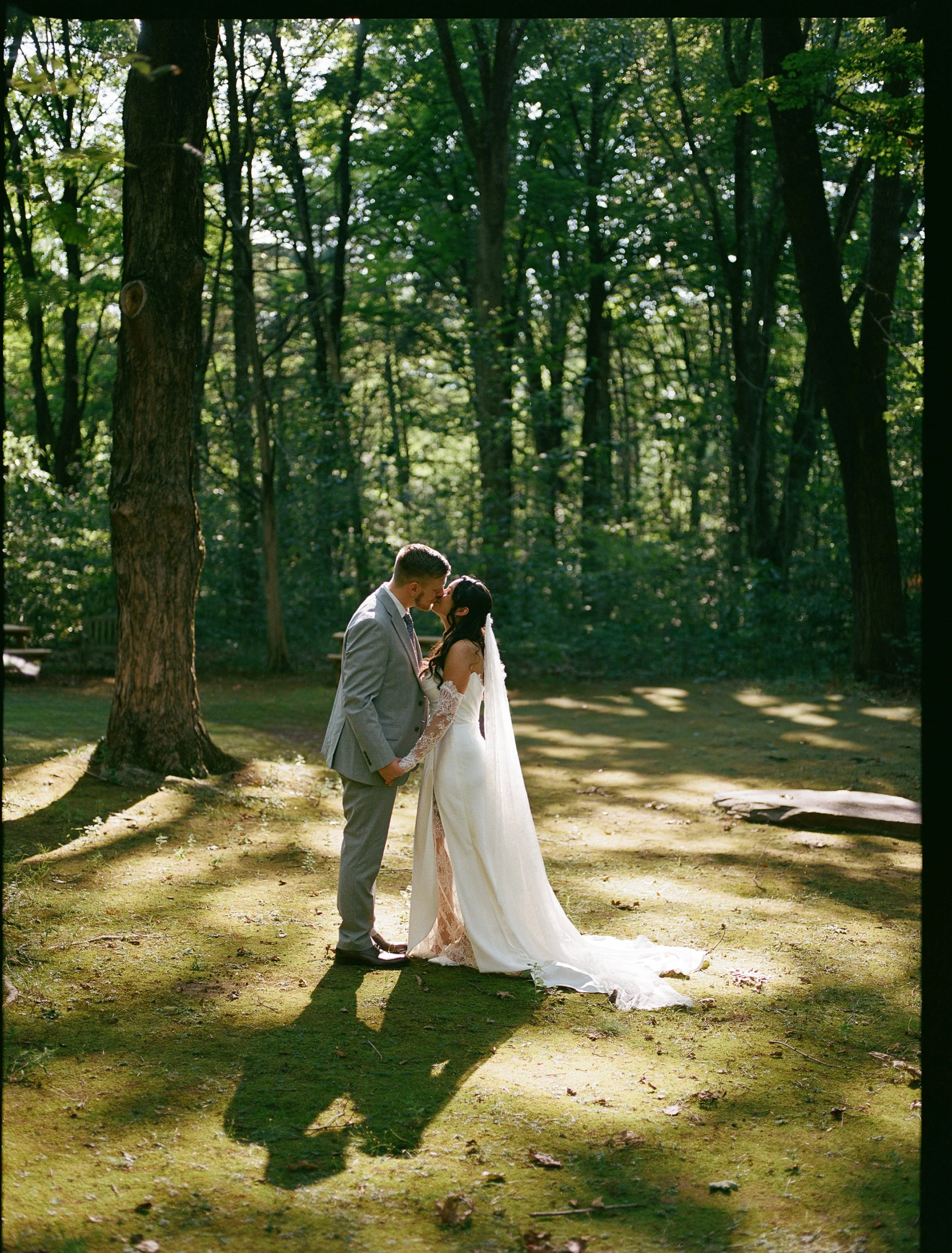 A couple in wedding attire sharing a kiss in a wooded outdoor setting with sunlight filtering through trees.