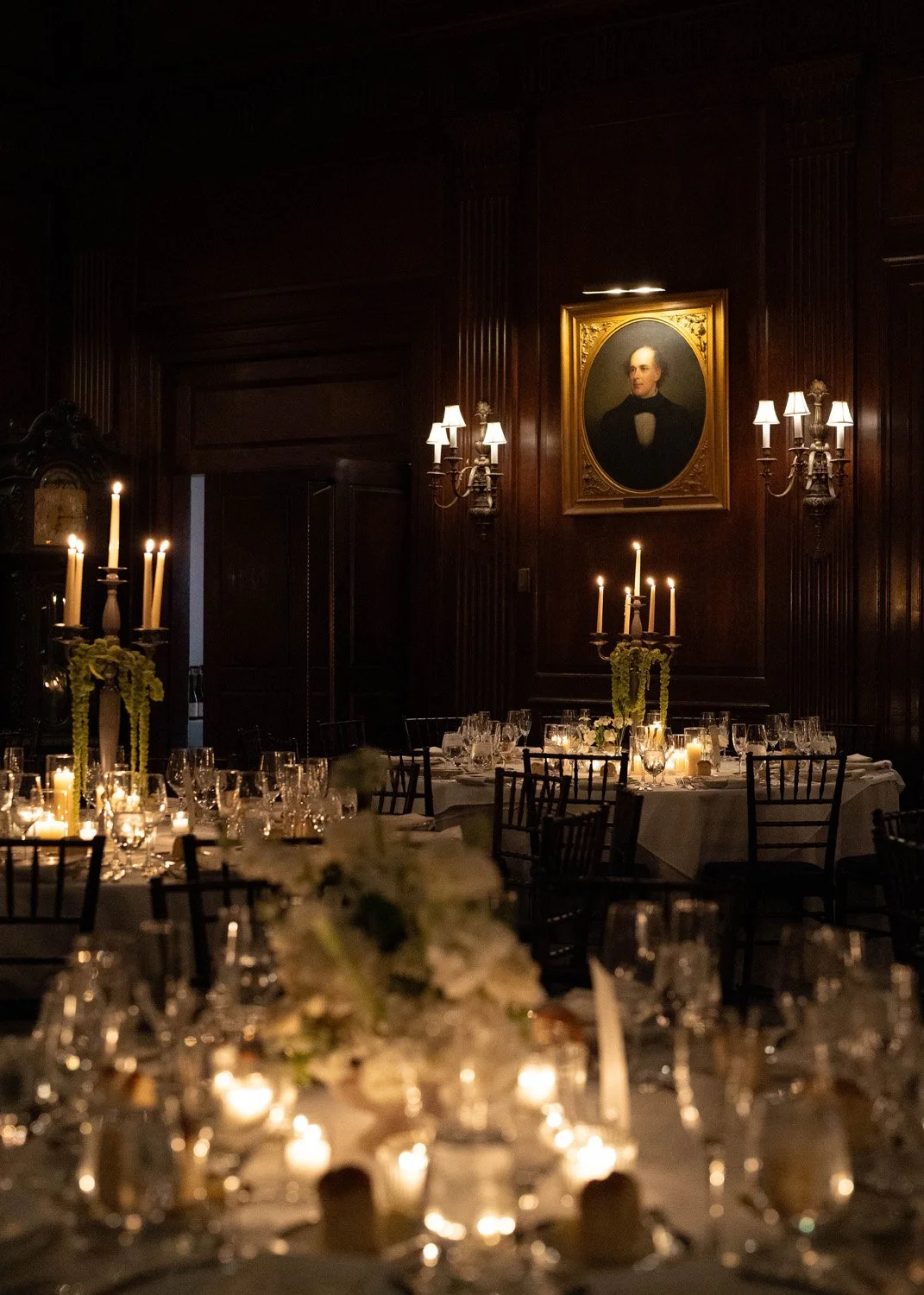 Elegant dining room with dark wood paneling, illuminated by wall sconces and candlelight, featuring a large portrait of a man in formal attire hanging on the wall.
