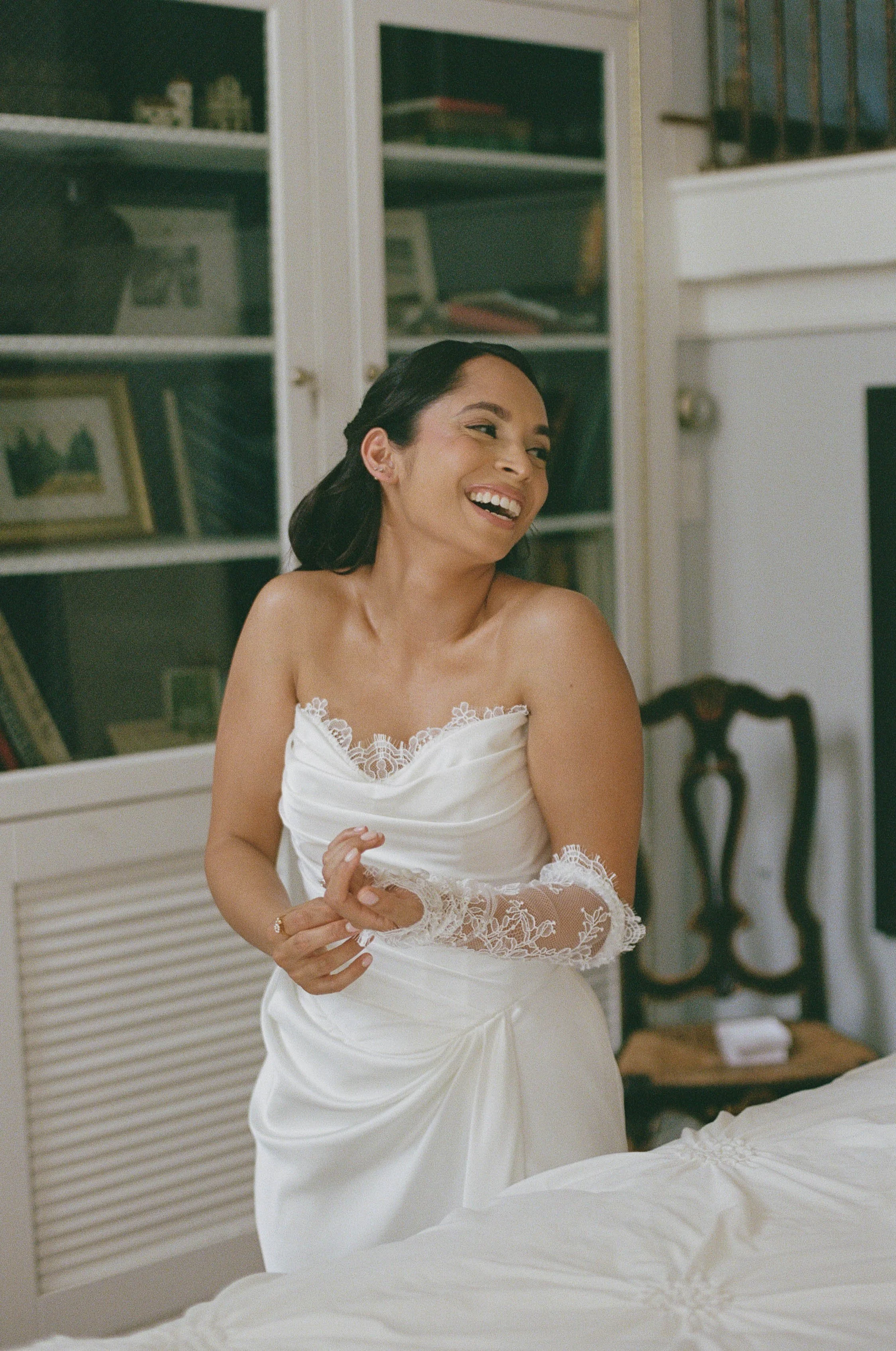 A woman in a white dress smiling and looking to the side in a room with bookshelves and a white bed.