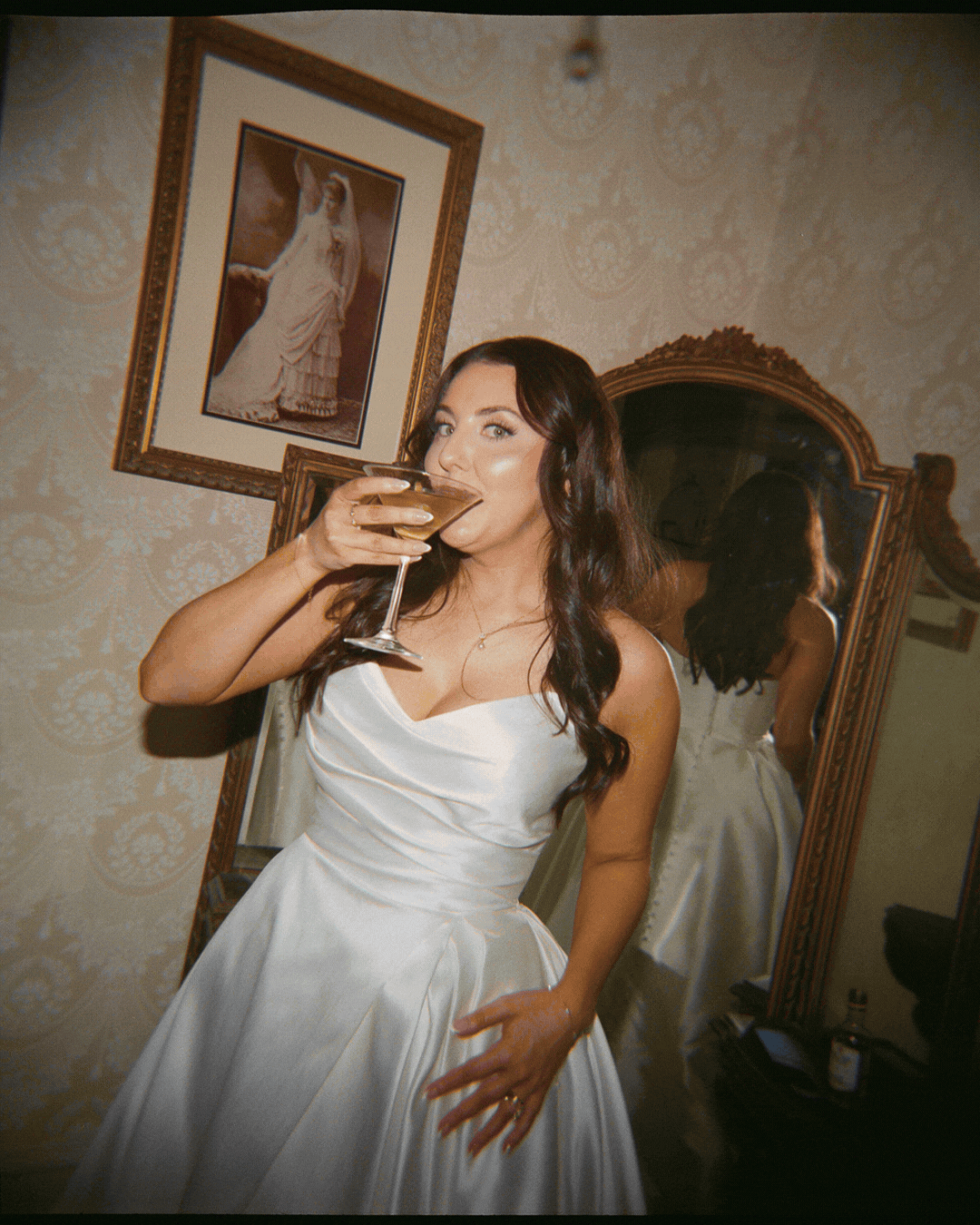 Woman in a white wedding dress drinking from a martini glass in a bedroom with an ornate mirror and framed artwork on the wall.