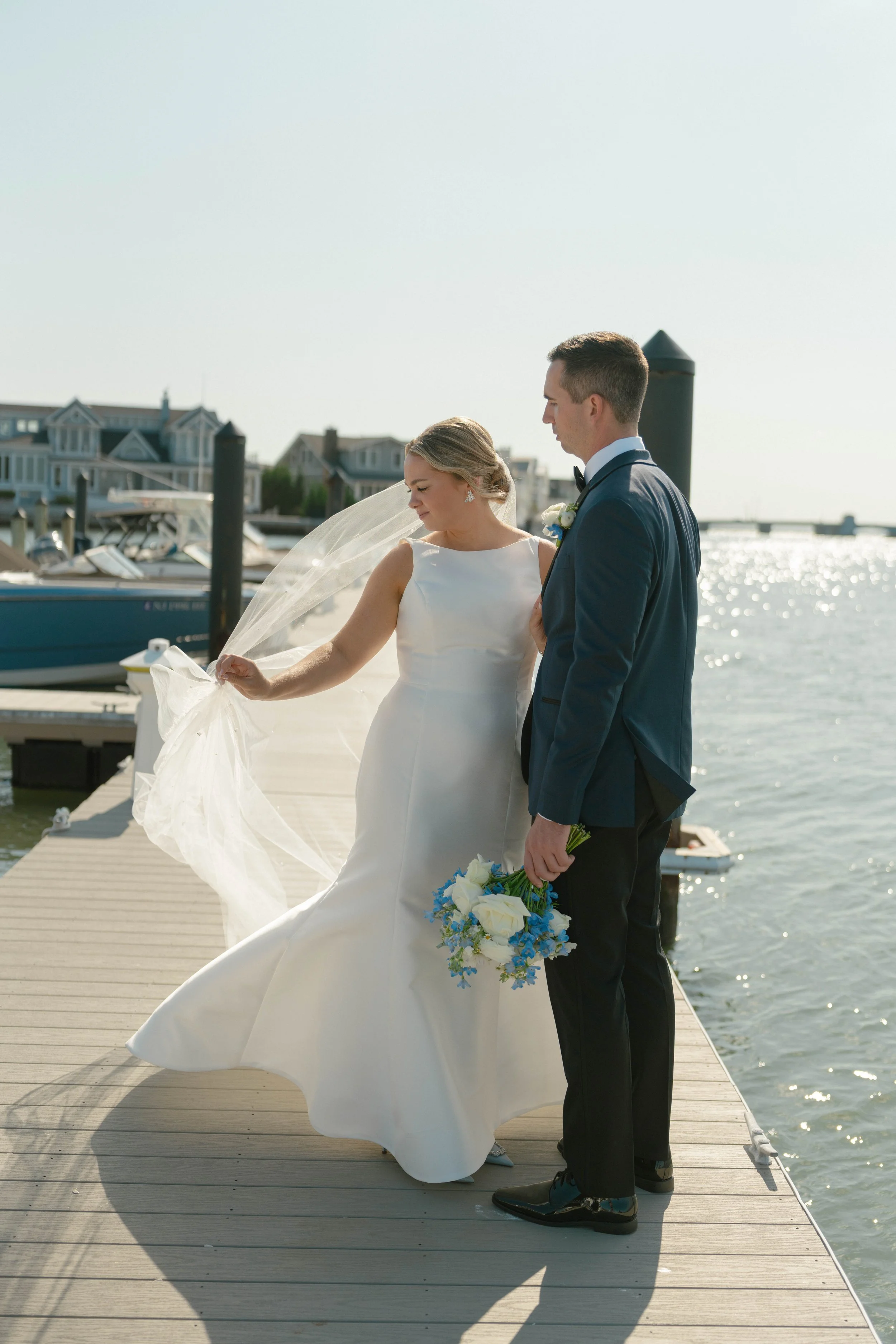 A bride and groom standing on a dock by the water, sharing a moment during their wedding celebration on a sunny day with boats and houses in the background.