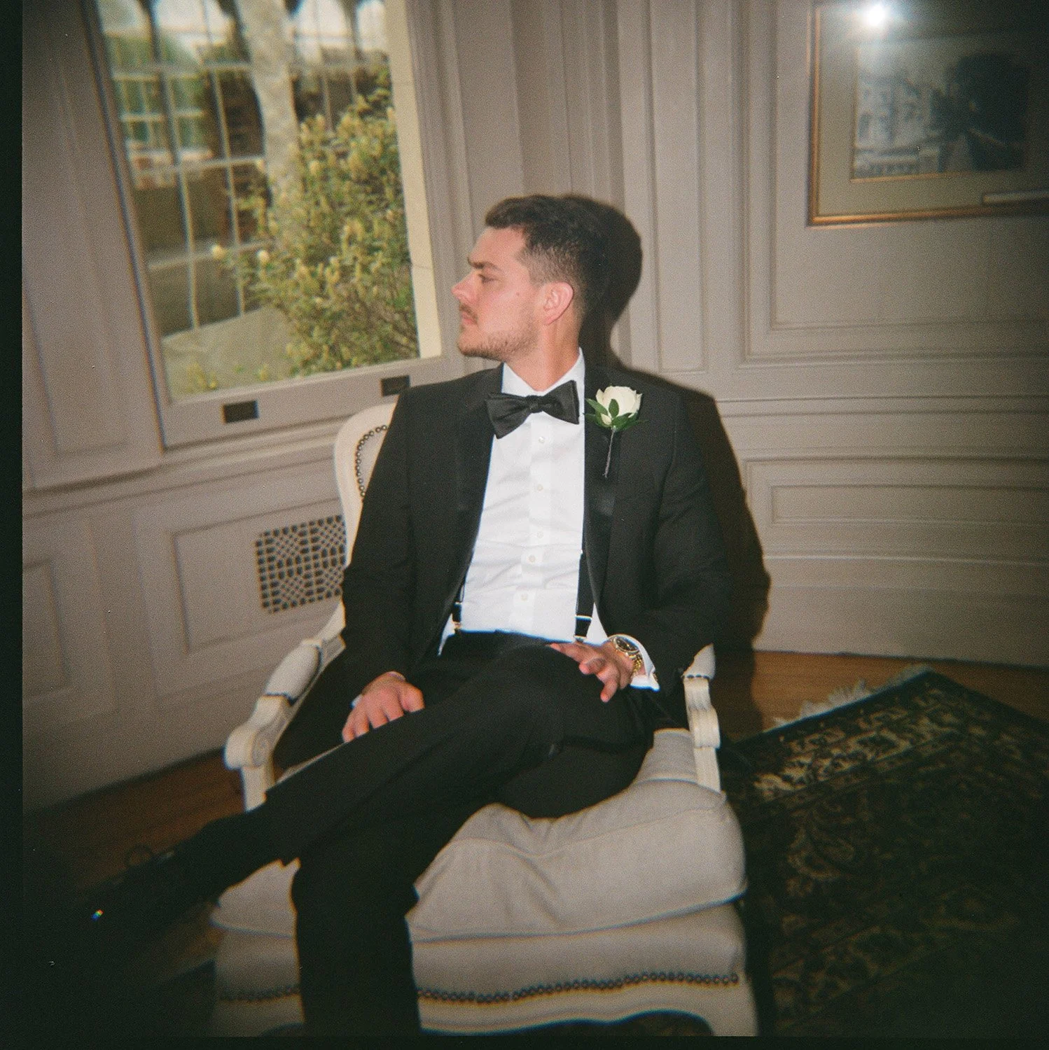 A man in a tuxedo with a white flower boutonniere sitting on a white chair inside a room with cream-colored walls and large windows, looking to the side.