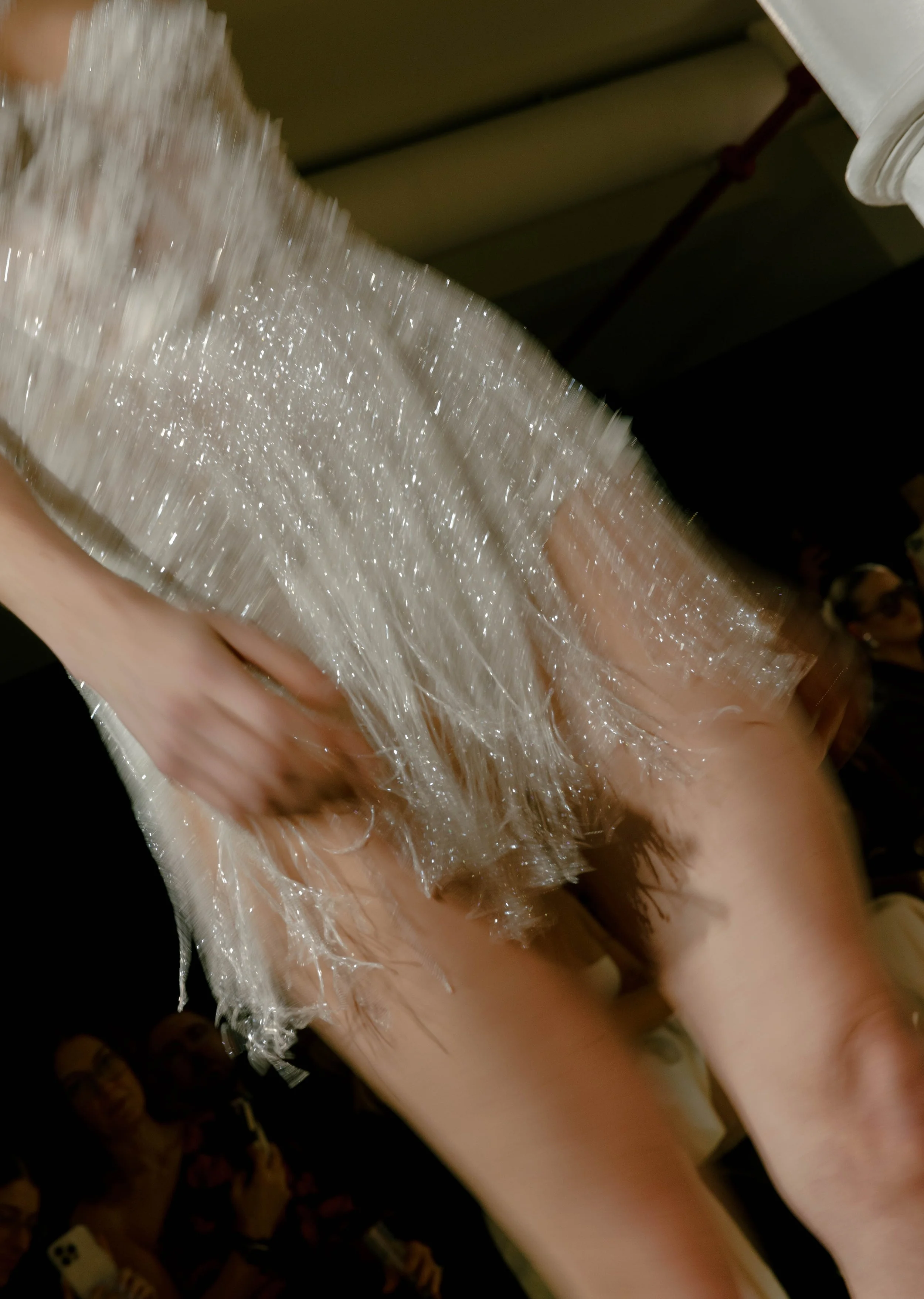 Close-up image of a person wearing a shiny, shimmering, transparent dress or outfit, possibly at a formal or special event, with an audience in the background.