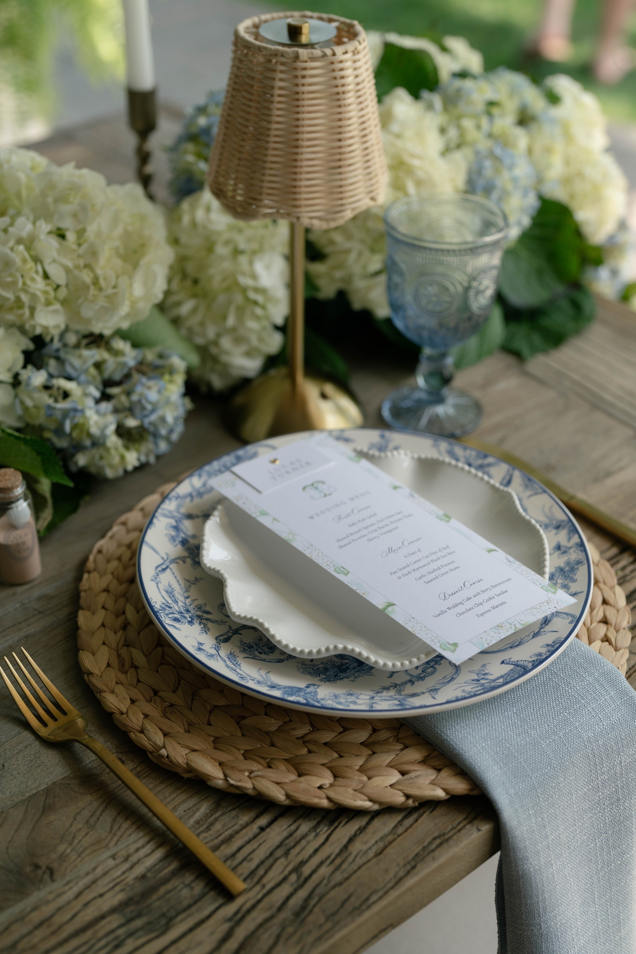 A rustic table setting featuring a woven placemat, a decorative blue and white plate, a white scalloped-edge dinner plate with a printed menu, a gold-colored fork, a gray napkin, surrounded by white hydrangea flowers, a small glass, a wicker lamp, a 