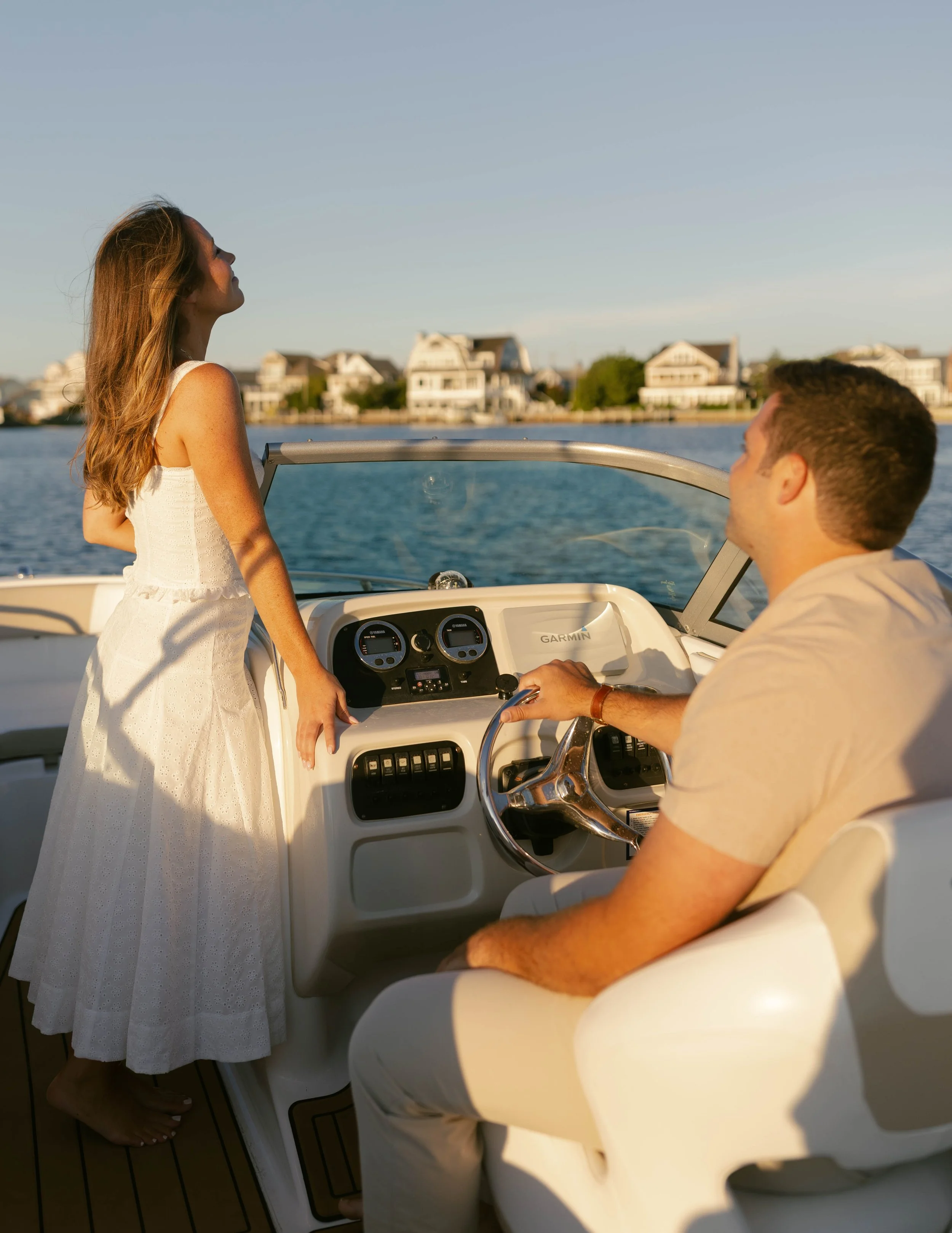 A woman in a white dress and a man in a beige shirt on a boat, with the woman standing and the man sitting at the helm, near a body of water and residential houses in the background during sunset.