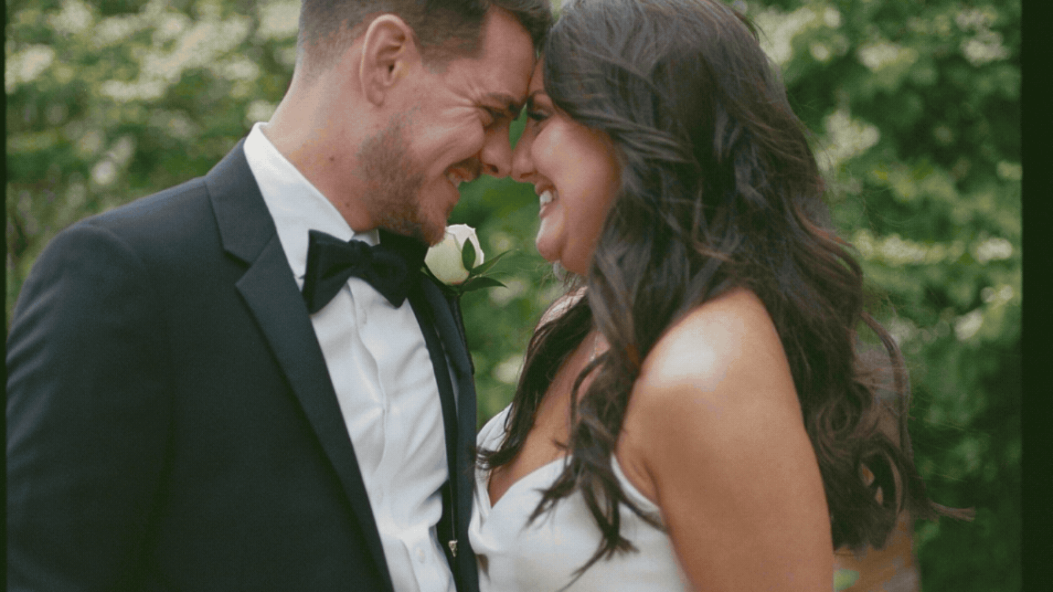 A bride and groom touching foreheads, smiling, in an outdoor setting with green foliage in the background.