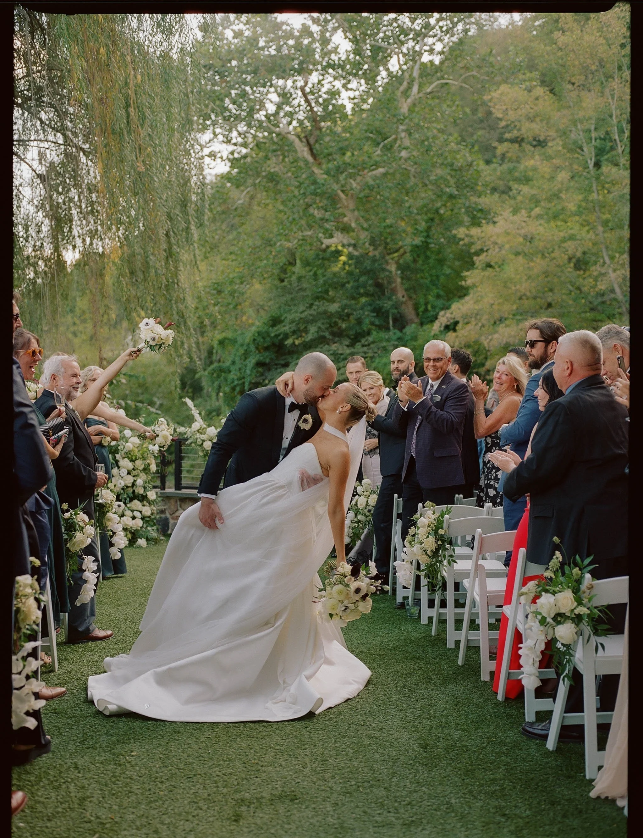 A bride and groom sharing a kiss at their outdoor wedding, with wedding guests clapping and smiling in the background, decorated with white flowers and greenery.