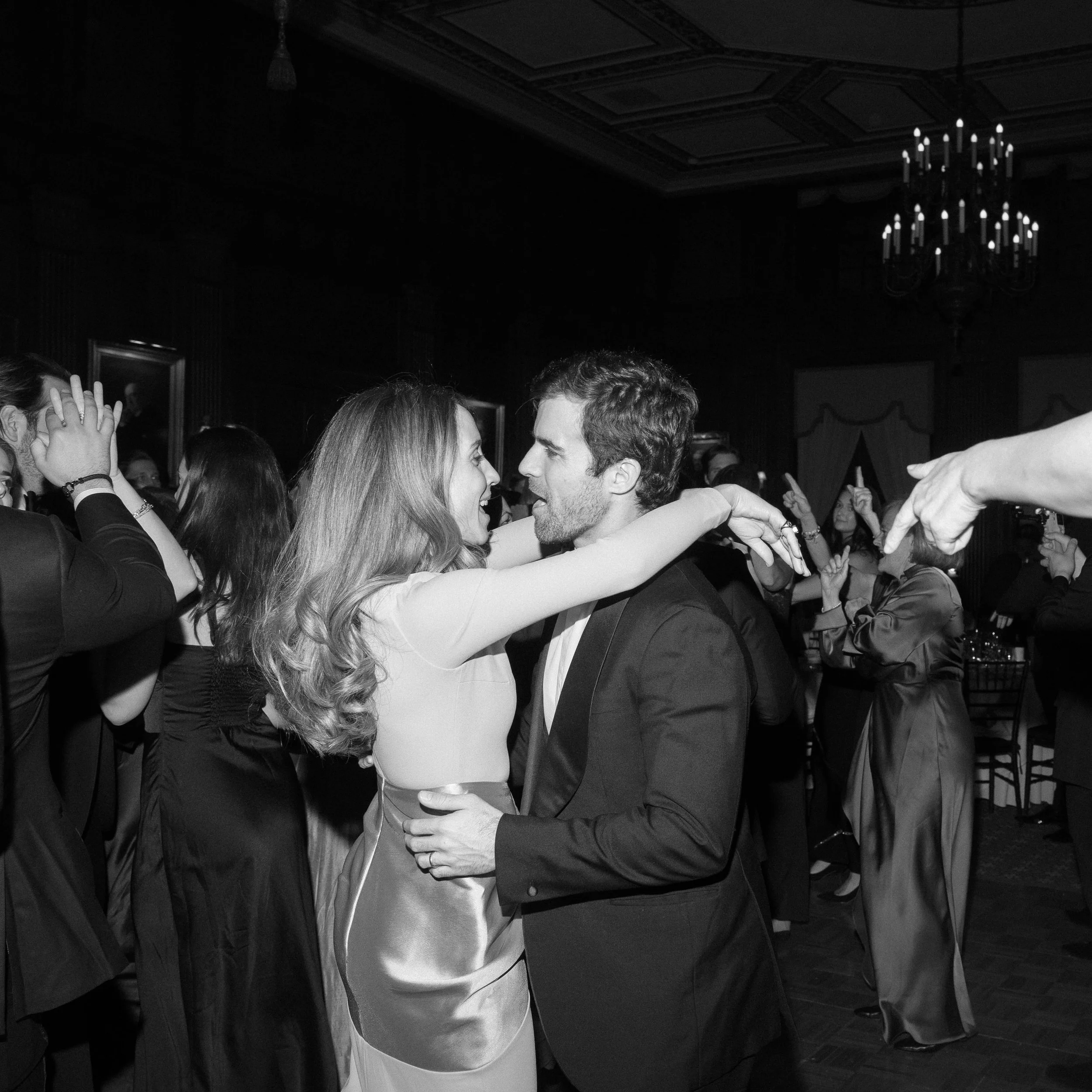 Couple dancing closely at a formal event, with other guests in the background and a chandelier hanging from the ceiling.