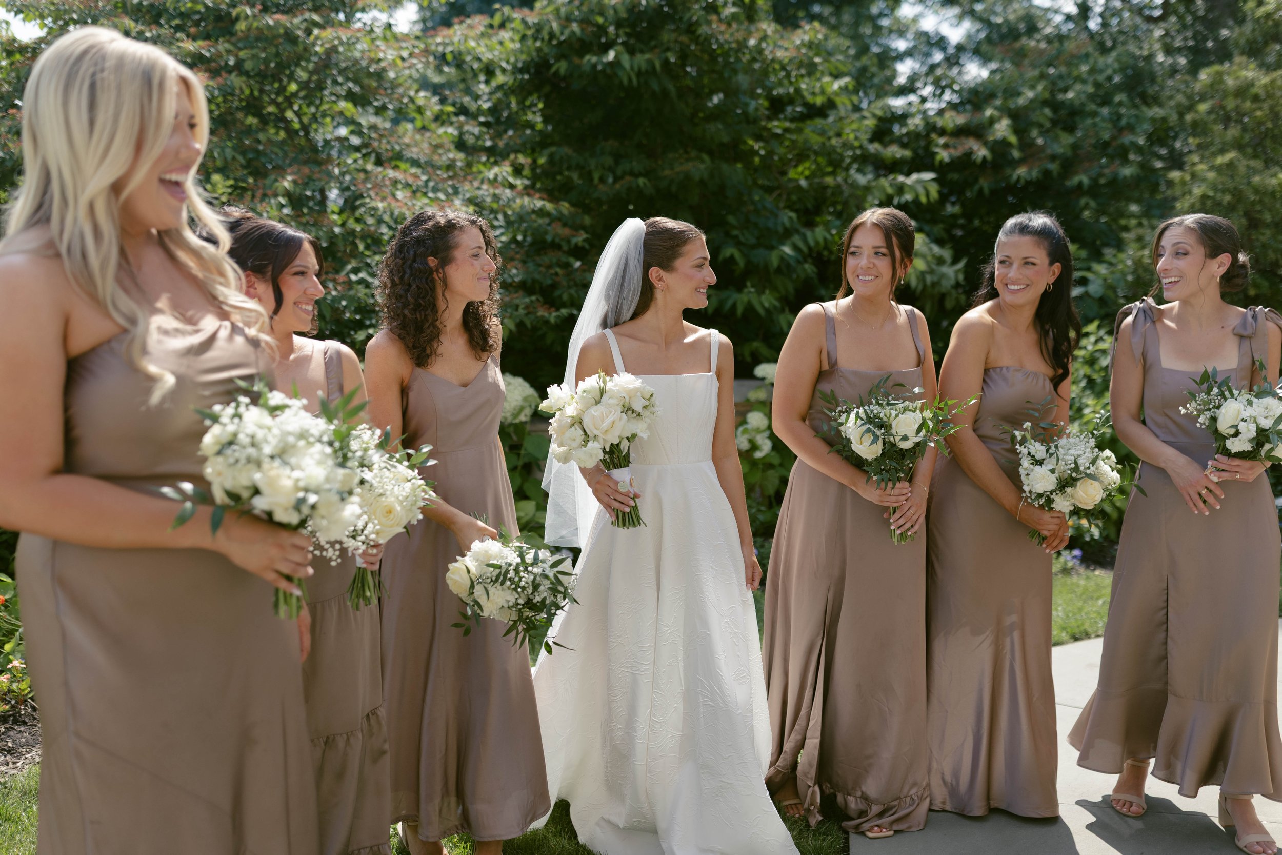 A bride in a white wedding dress and veil standing among bridesmaids in taupe dresses, all holding white bouquets, outdoors with lush greenery in the background.