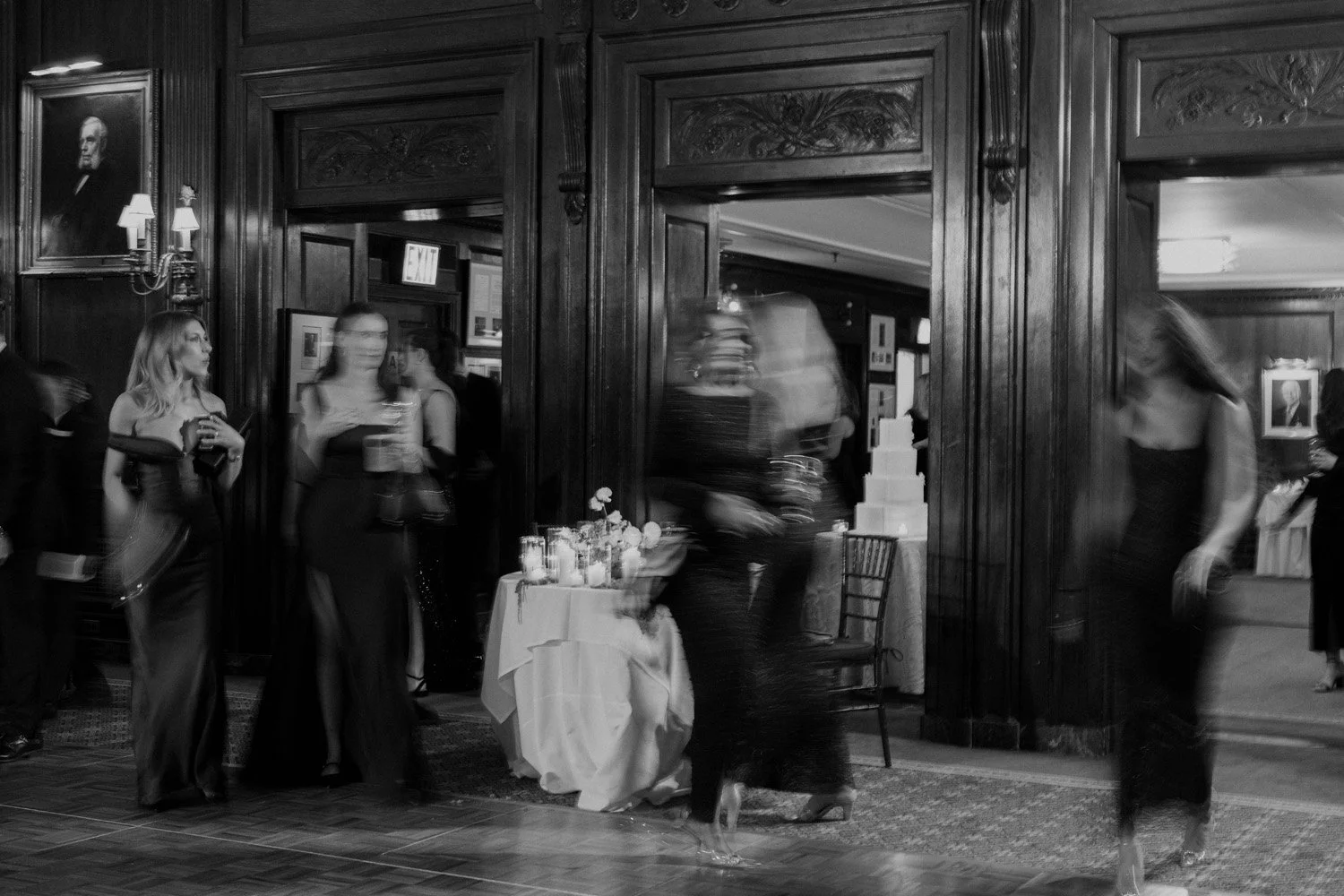 People socializing at wedding in an elegant, wood-paneled room with framed portraits on the wall, a table with candles and flowers, and a staircase in the background.