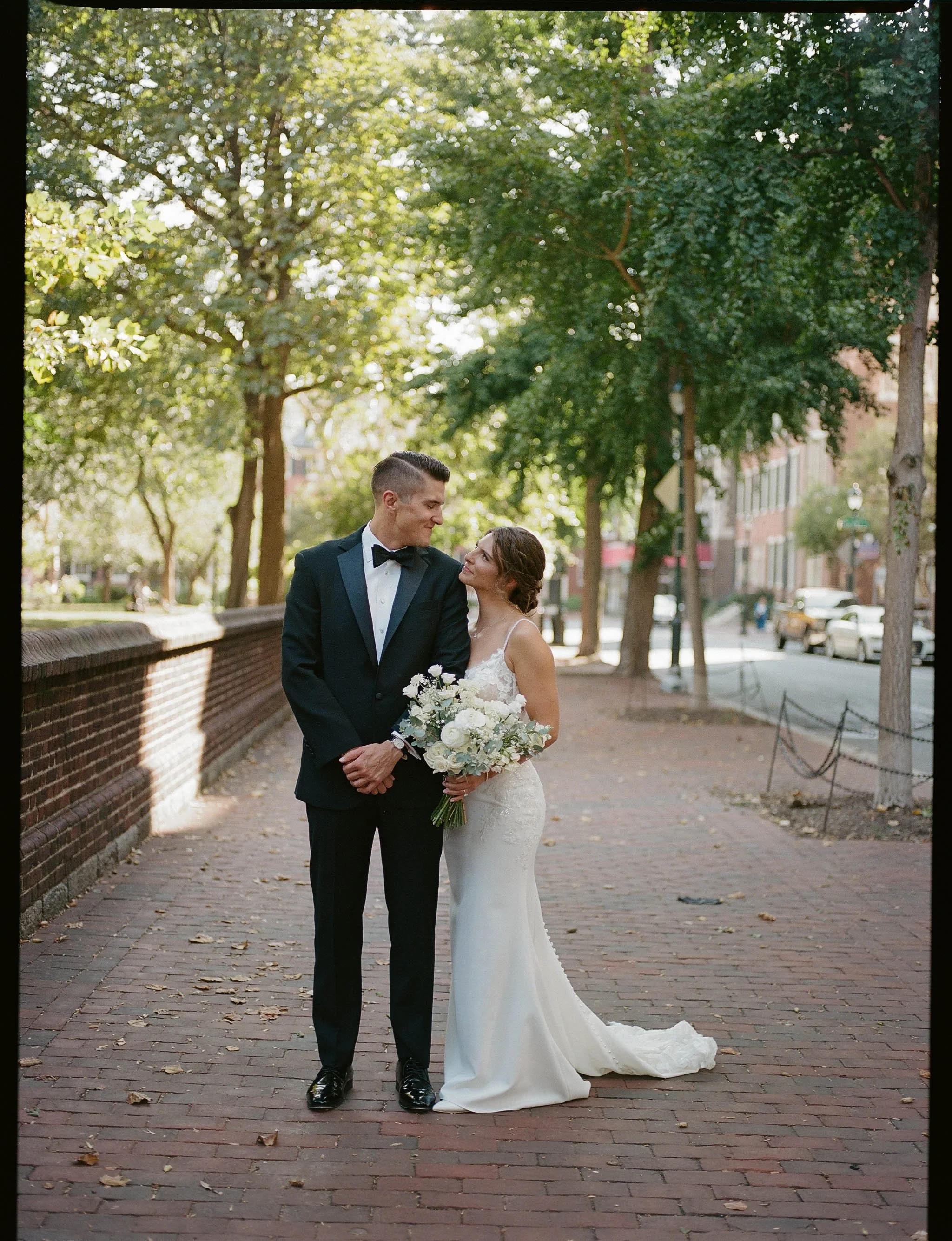A newlywed couple standing close together on a brick sidewalk in a park, surrounded by tall trees and lush green foliage. The groom in a black tuxedo with a bow tie looks at the bride, who is in a white wedding gown holding a bouquet of white flowers