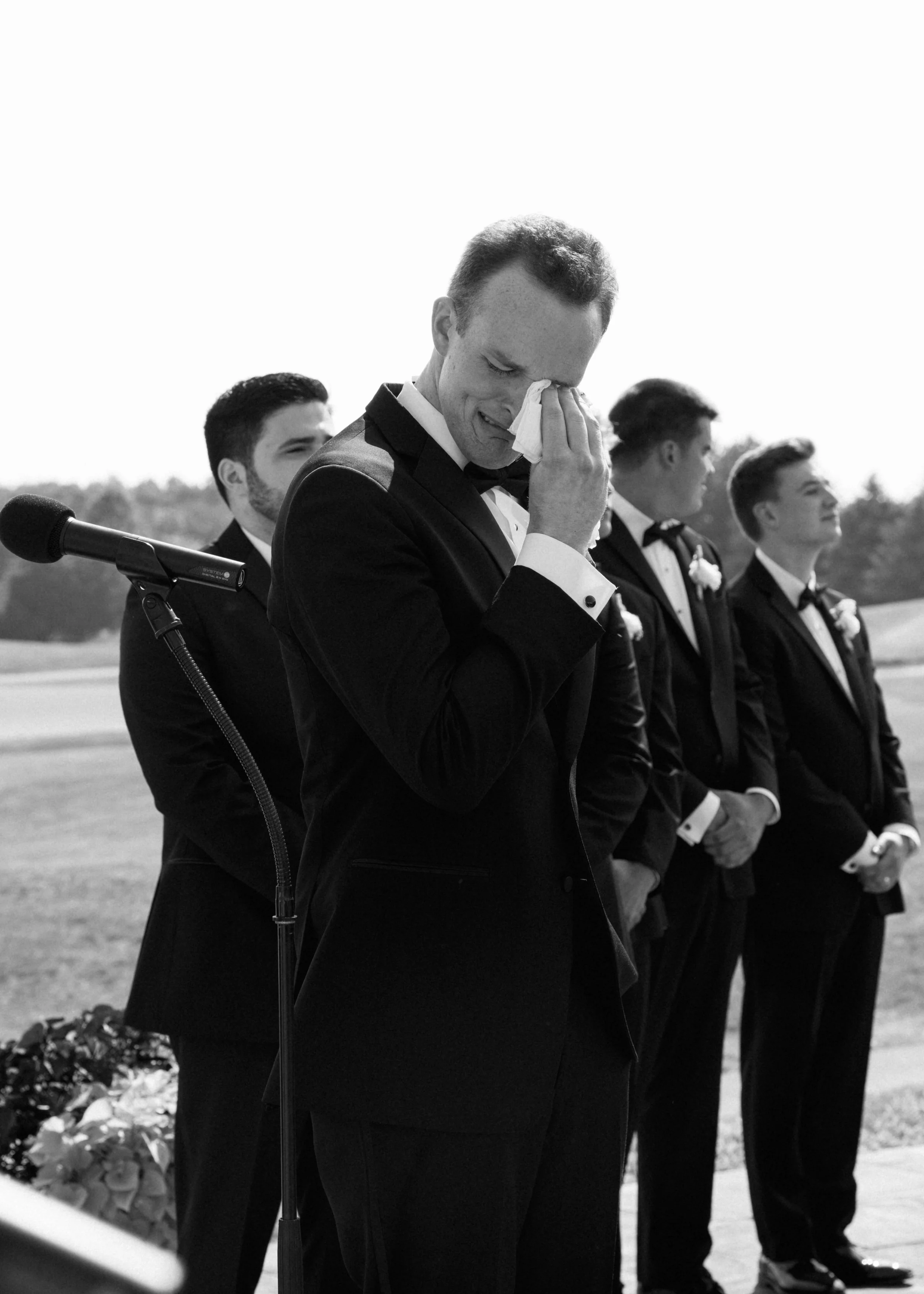 A grieving man in a tuxedo wipes his face with a tissue at an outdoor memorial service, surrounded by men in tuxedos, with flowers on the ground nearby.