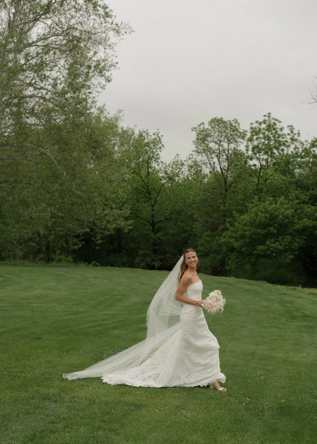A bride in a white wedding dress holding a bouquet of flowers, standing on a green lawn with trees in the background on a cloudy day.