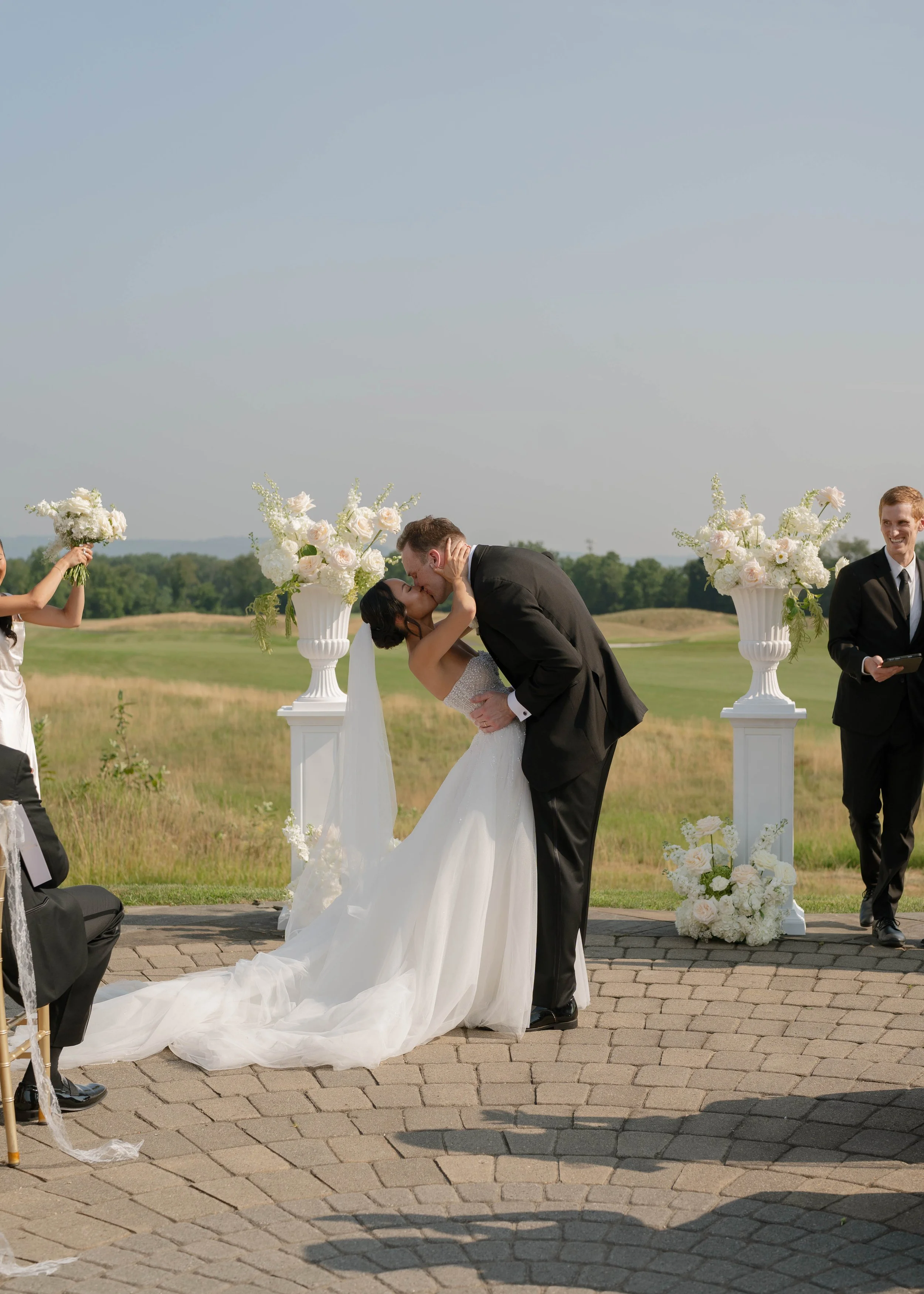 A couple kissing during an outdoor wedding ceremony with officials in formal attire, floral decorations, and a scenic landscape in the background.