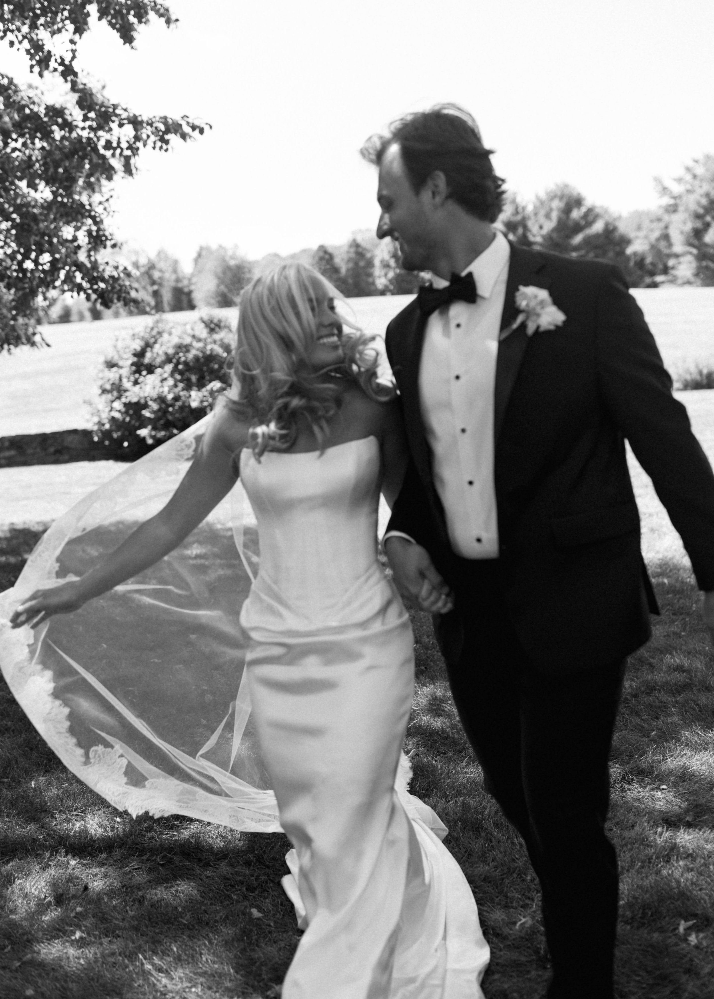 A happy couple wearing wedding attire, standing outdoors on grass, smiling at each other in a black-and-white photo.