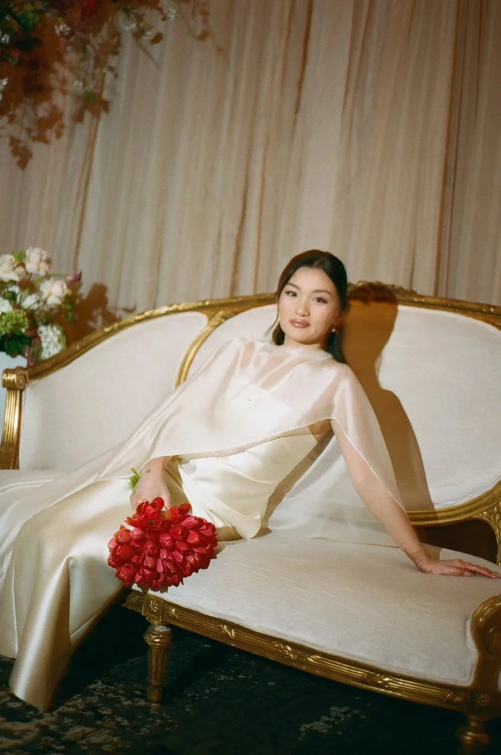 A woman in a white dress sitting on a vintage sofa, holding a bouquet of red flowers with a floral arrangement in the background.