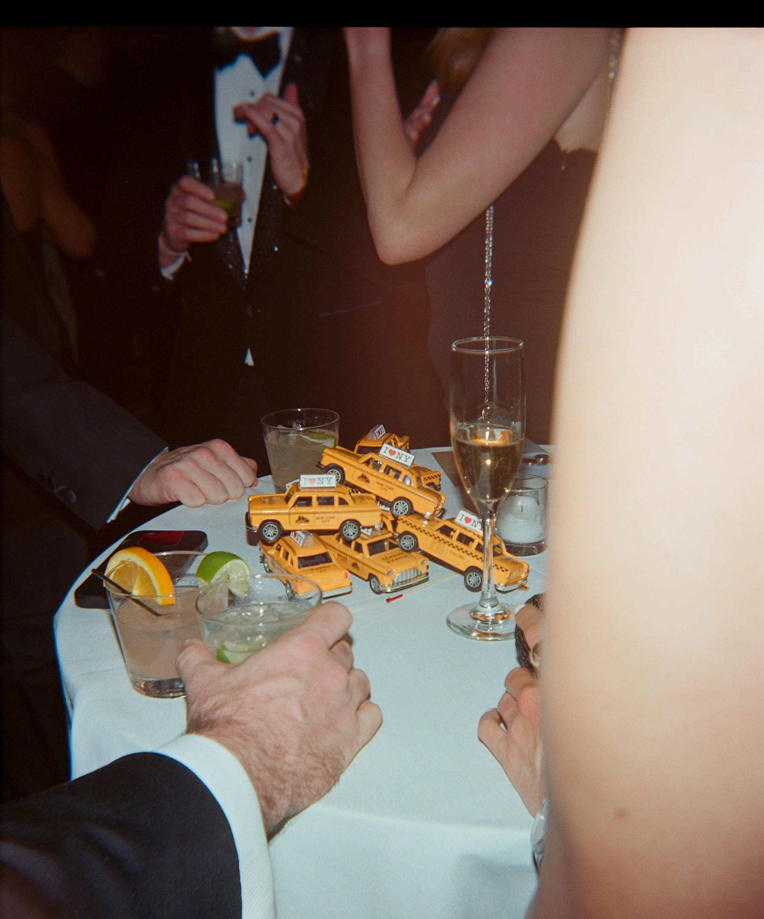 People celebrating at a wedding reception, holding drinks and toasting, with New York taxi cab toys and 'I ♥ NY' buttons on the table.