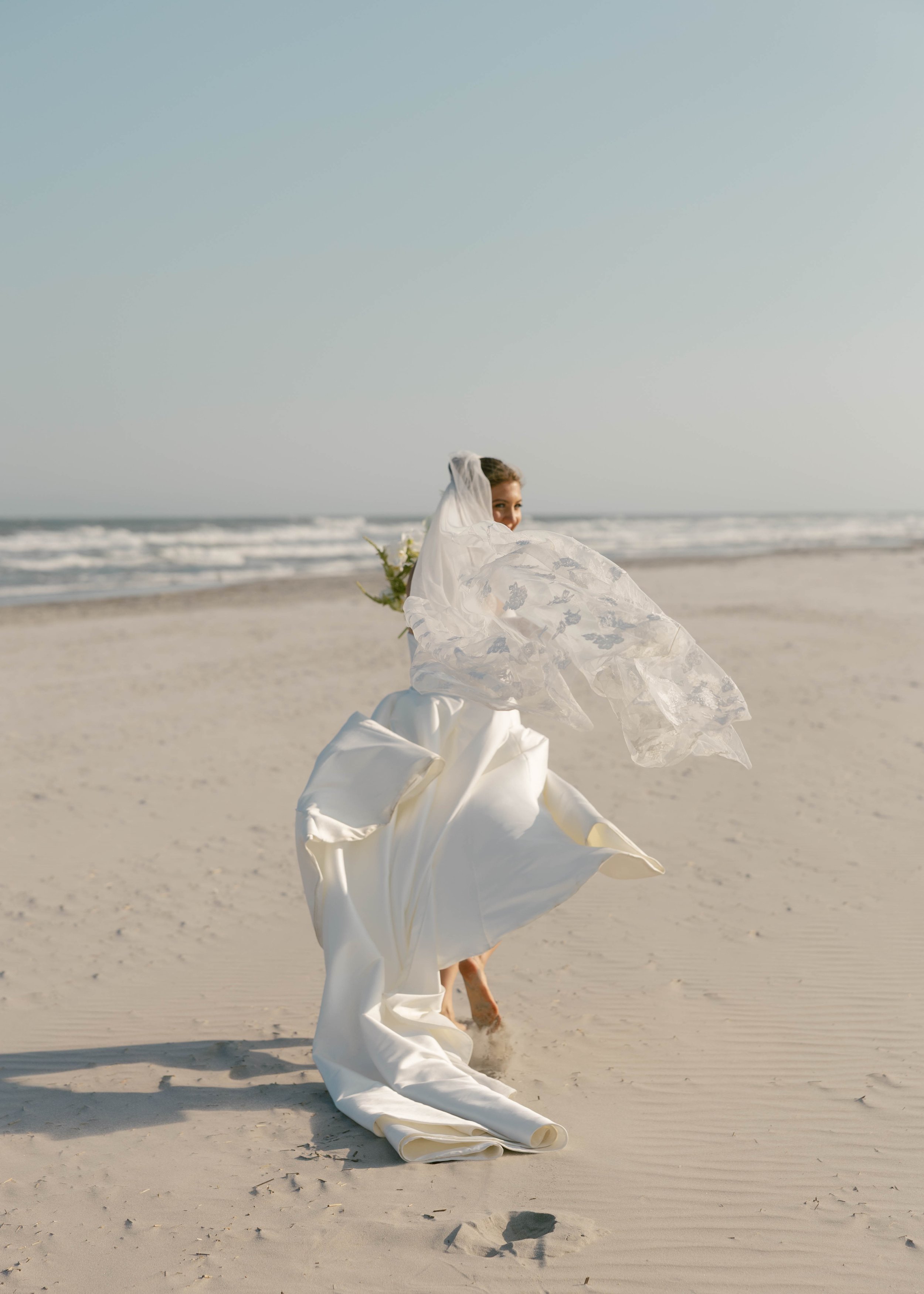 A woman in a white wedding dress running on the beach with the ocean in the background, her veil flowing behind her.