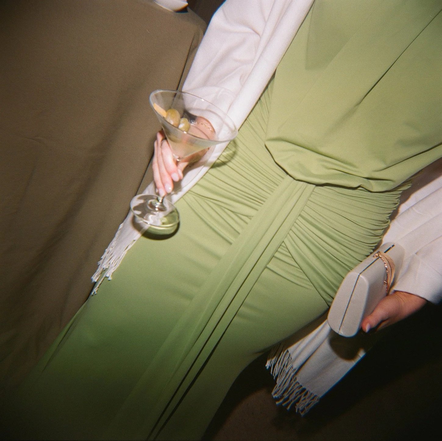 Close-up of a woman in a green dress sitting on a beige chair, holding a martini glass with olives and a cocktail.