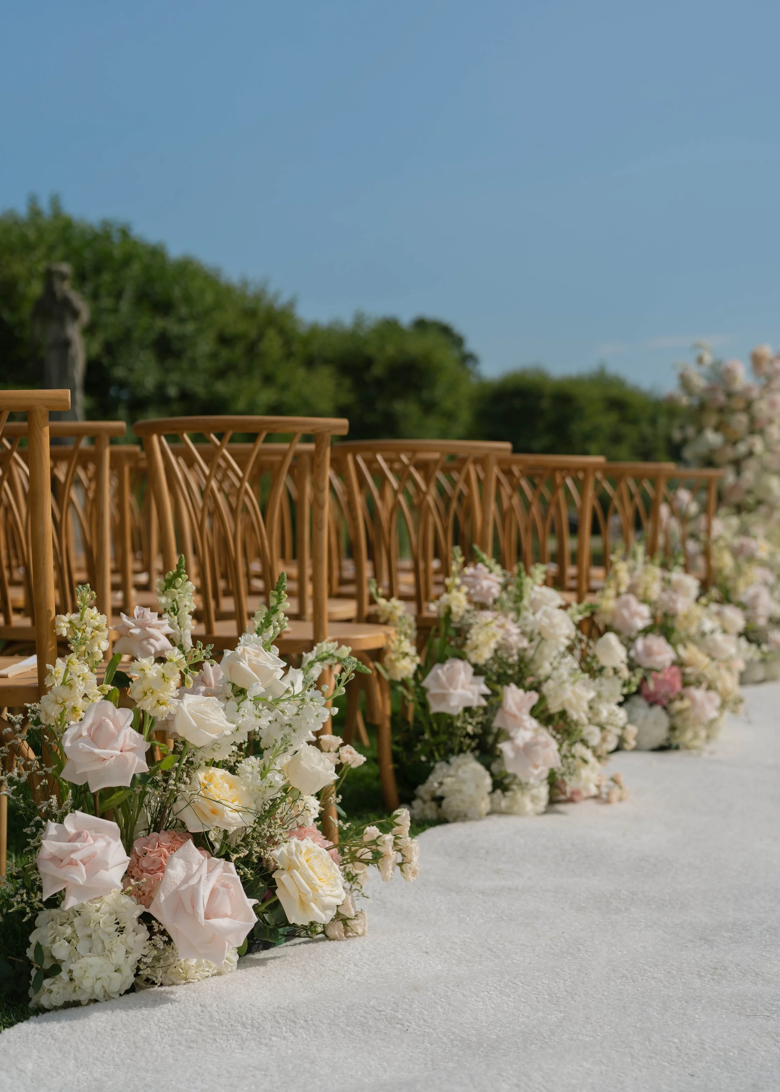 Rows of wooden chairs at an outdoor wedding ceremony, decorated with white and pale pink flowers along the aisle, with green trees and clear blue sky in the background.
