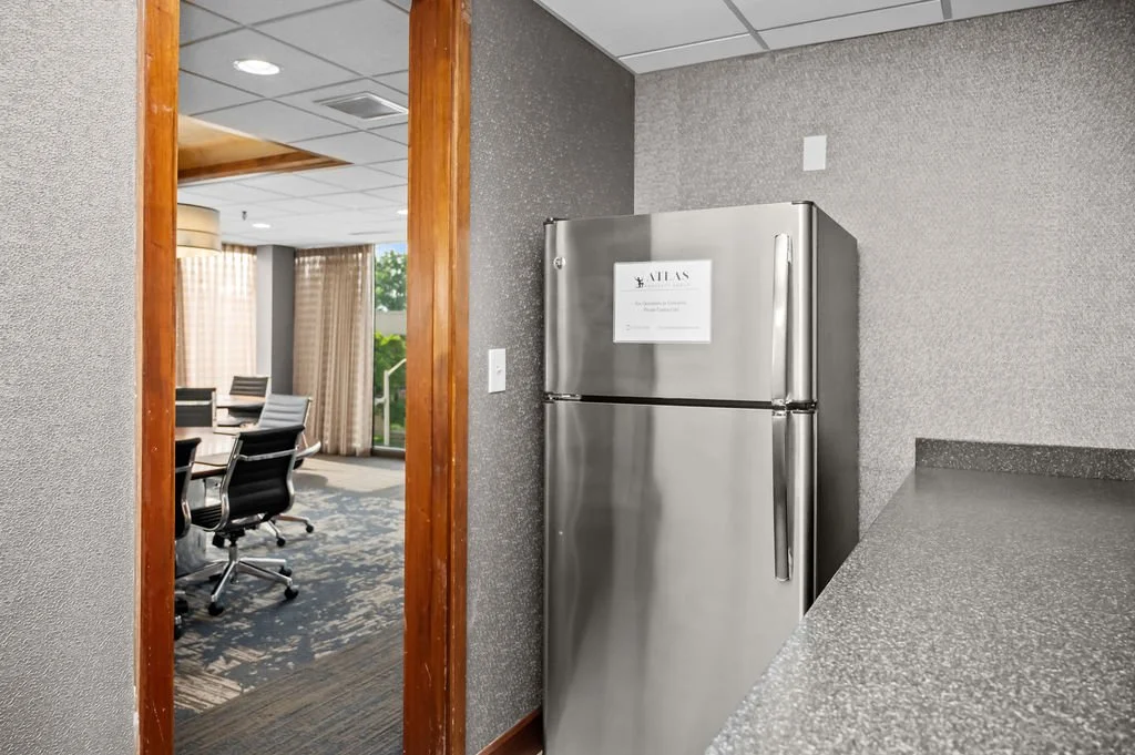 A stainless steel refrigerator in a conference room with chairs and a table visible through a doorway.