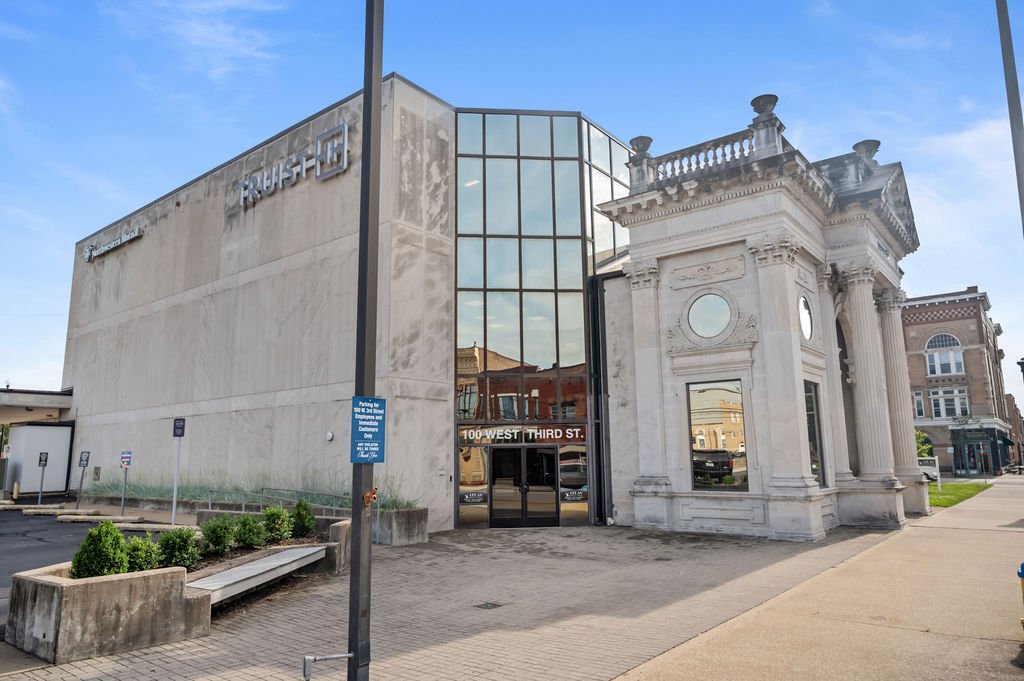 A building with modern architecture on the left and classical architecture on the right, with a glass entrance door in the center. The address '100 West Third St.' is visible above the door. There is a parking sign and a small bench with shrubs in front of the building.