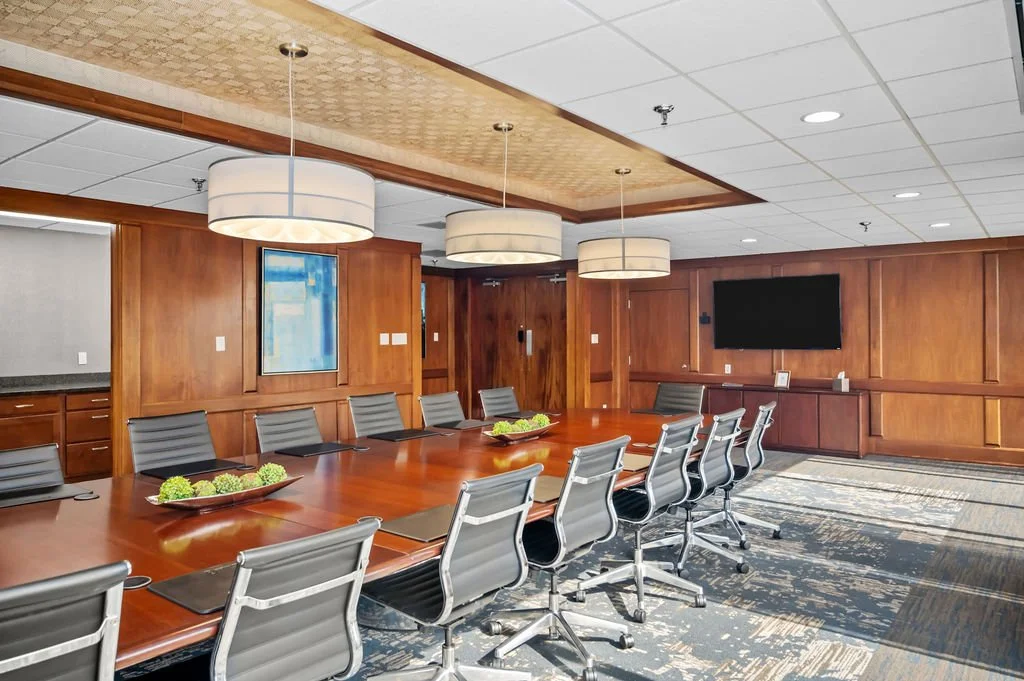 A conference room with a long wooden table and multiple gray office chairs. The room has wood-paneled walls, a wall-mounted TV, and hanging ceiling lights. There are decorative plates with green plants on the table, and artwork on the wall.