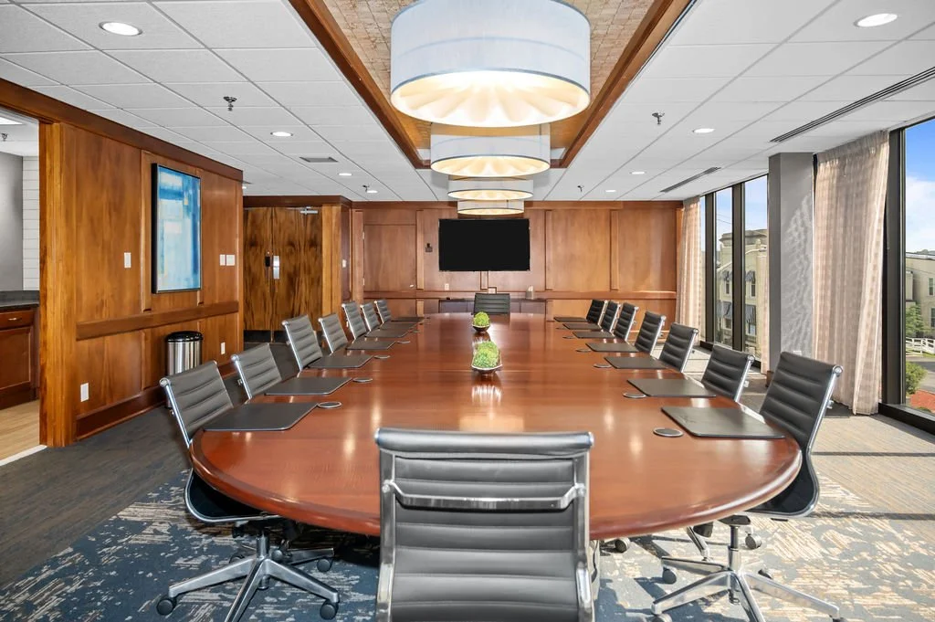 A modern conference room with a long wooden table, black leather chairs, and large windows with curtains, with a view outside.