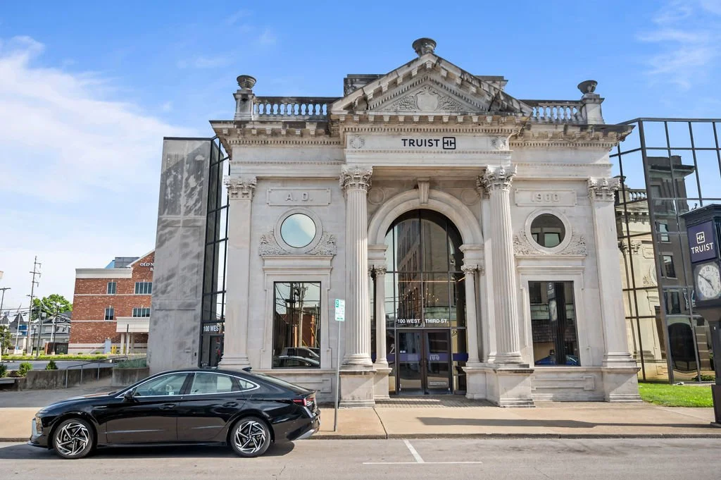 A historic concrete building with classical architecture style, featuring pillars, decorative details, and a pediment. A black sedan is parked in front of the building, and a clear blue sky is above.