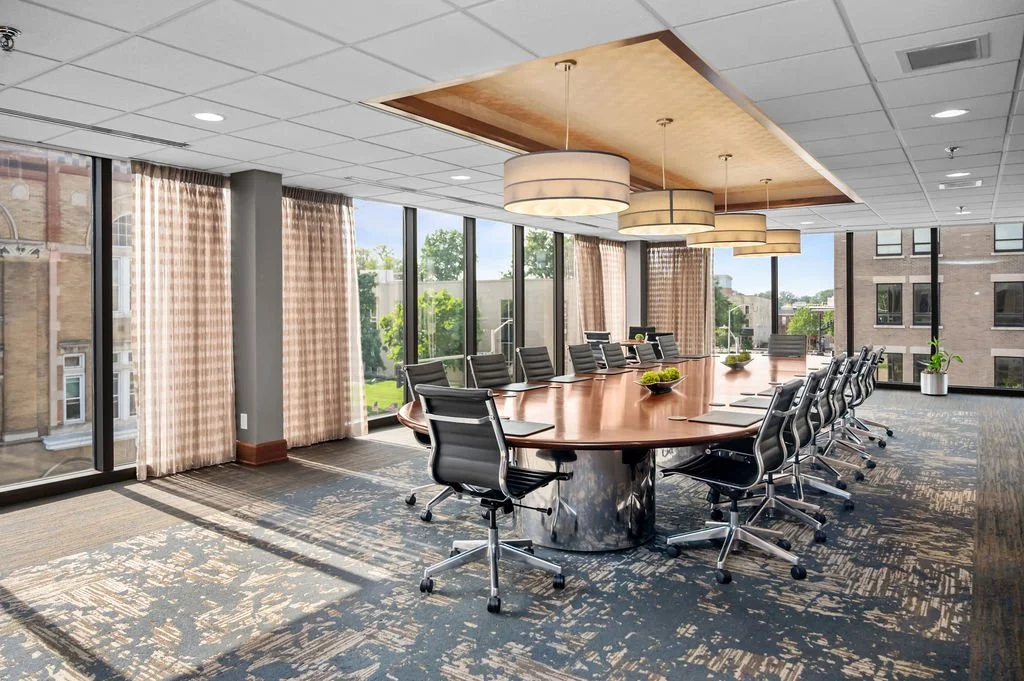 A modern conference room with a large oval wooden table, surrounded by black swivel chairs. The room has floor-to-ceiling windows with beige checkered curtains, allowing natural light. There are ceiling-mounted circular pendant lights and a wooden ceiling feature. Small green plants are on the table and window sill, with a view of neighboring buildings and trees outside.