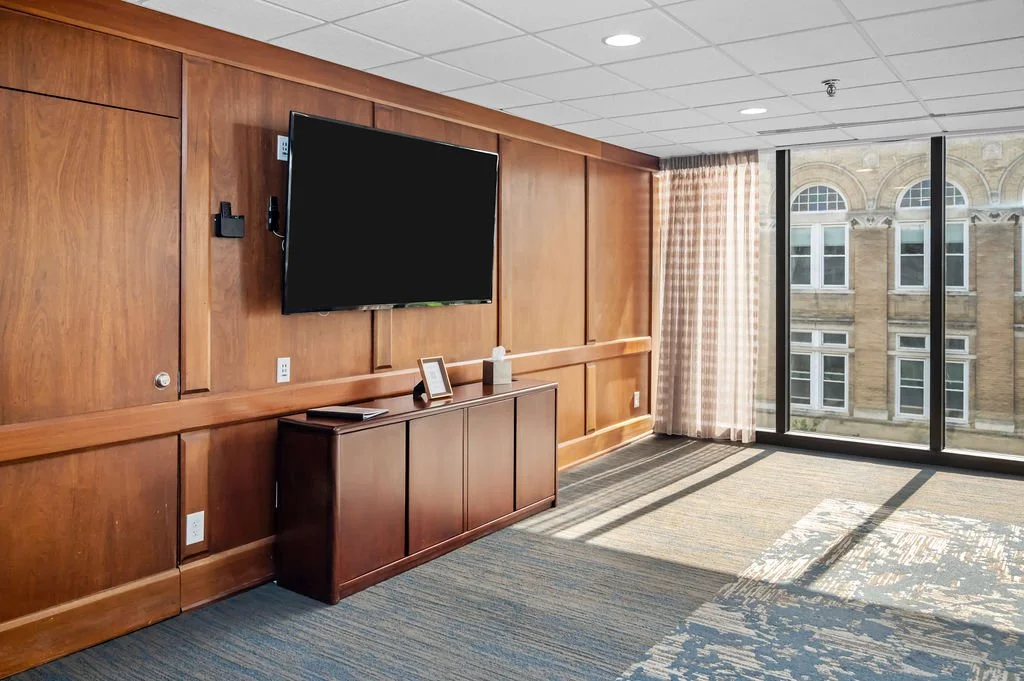 Room with wooden wall paneling, large flat-screen TV mounted on the wall, a cabinet below with a framed photo or sign, and windows with checkered curtains letting in natural light.