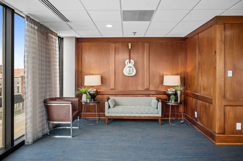 A cozy corner in a room with wood-paneled walls, featuring a small gray couch, two side tables with lamps, a brown armchair, and a white guitar mounted on the wall.