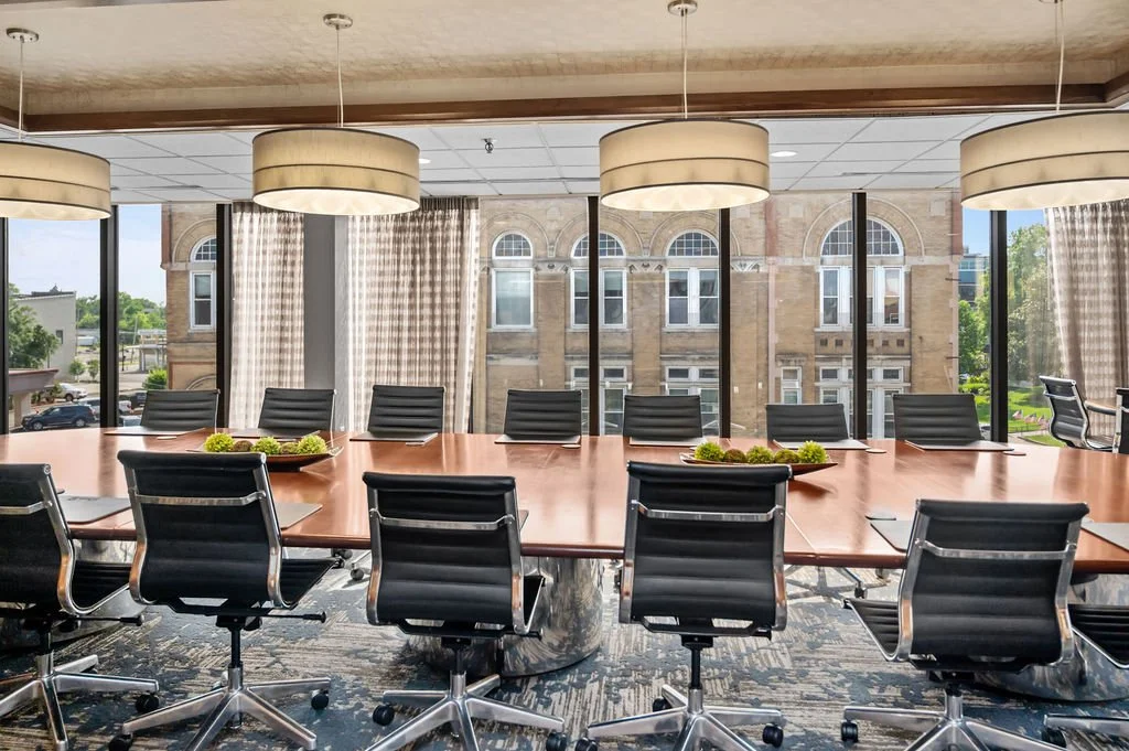 A modern conference room with a large wooden table and nine black leather chairs. The room has large windows with curtains, providing a view of buildings and green trees outside. Suspended ceiling lights hang above the table.