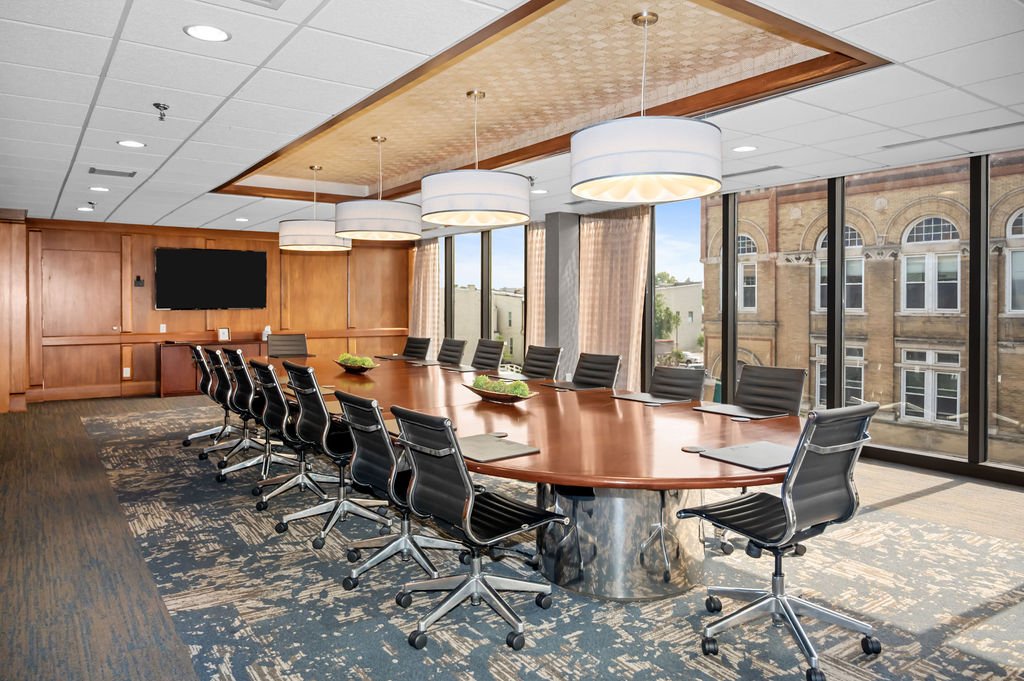 Empty modern conference room with a large oval wooden table, black chairs, pendant lights, and large windows with city view.
